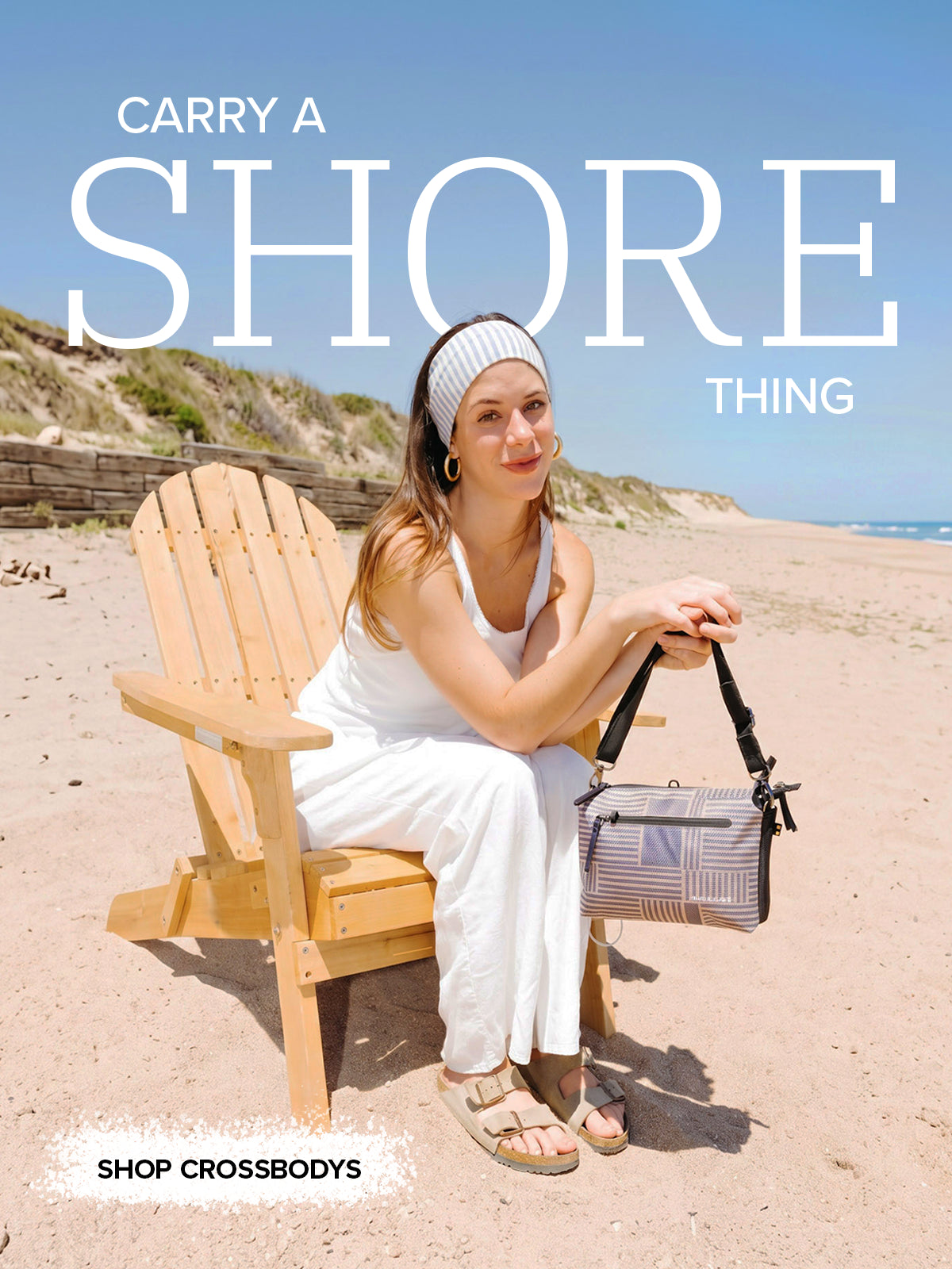 A woman sits on a wooden Adirondack chair on a sandy beach, wearing a white dress and sandals and holding a patterned Crossbodies handbag. Large white text at the top reads 'CARRY A SHORE THING' and smaller text at the bottom reads 'SHOP CROSSBODIES'.