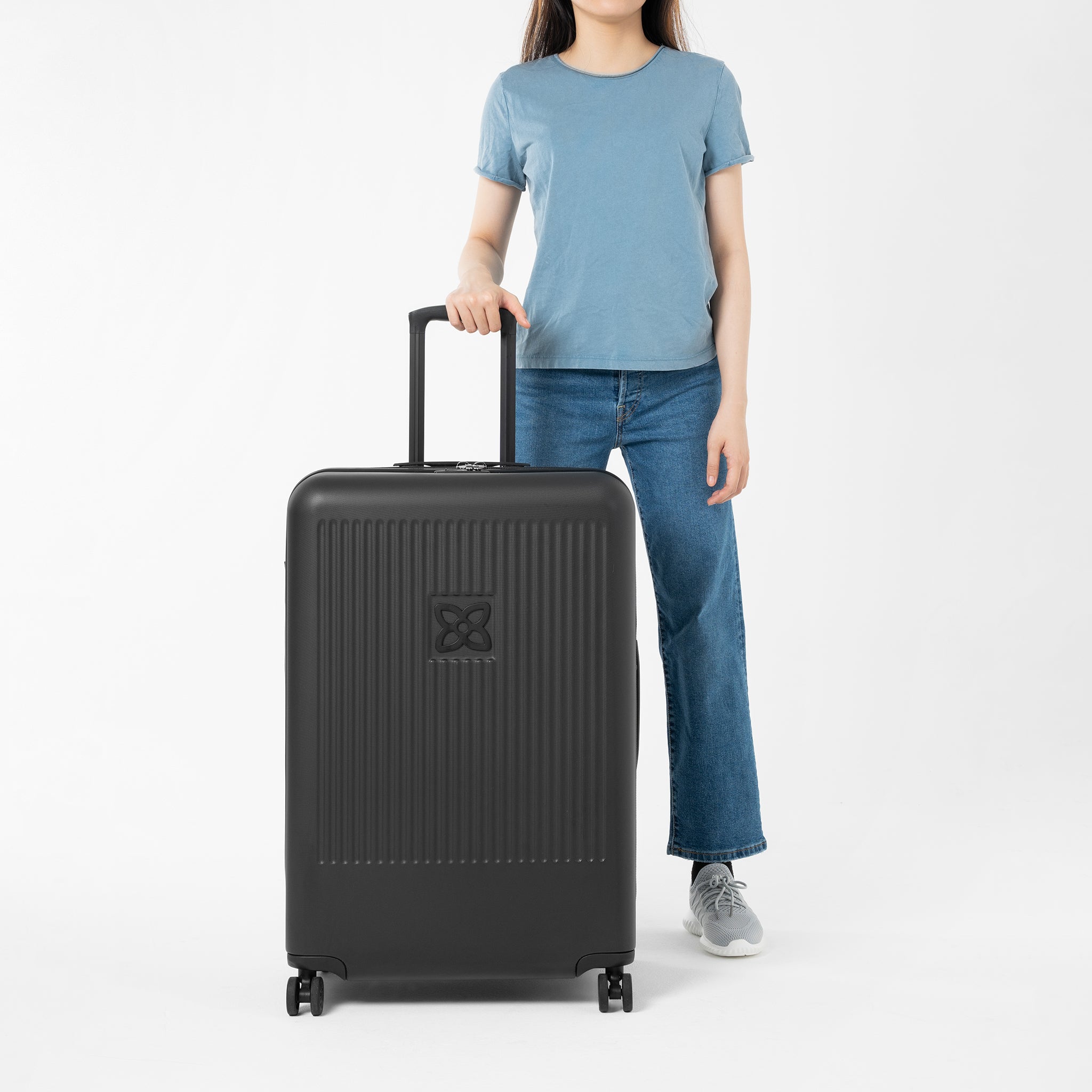 A model stands with one hand on the suitcase handle of Sherpani hard-shell luggage, the Meridian in Black. 
