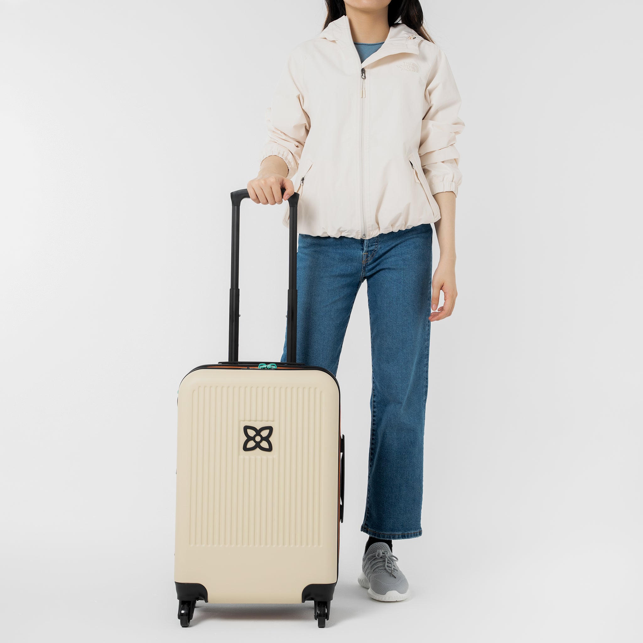 A model stands with one hand on the suitcase handle of Sherpani hard-shell carry-on suitcase, the Meridian in Bluff. 