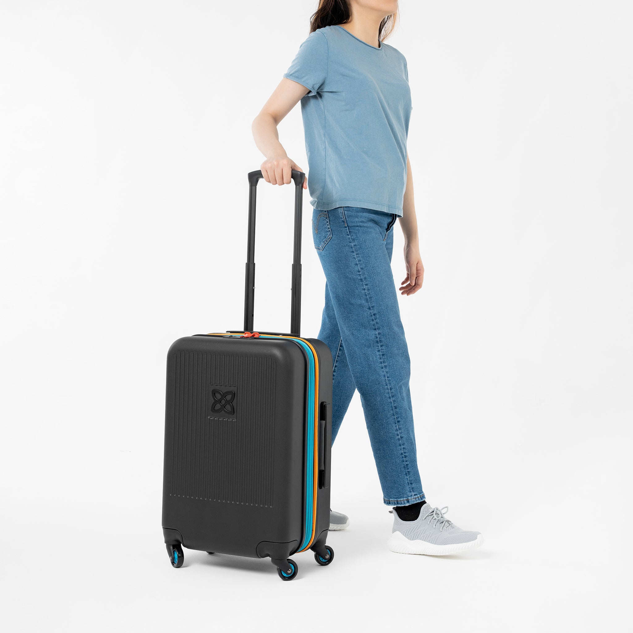 A model stands with one hand on the suitcase handle of Sherpani hard-shell carry-on suitcase, the Meridian in Chromatic. 