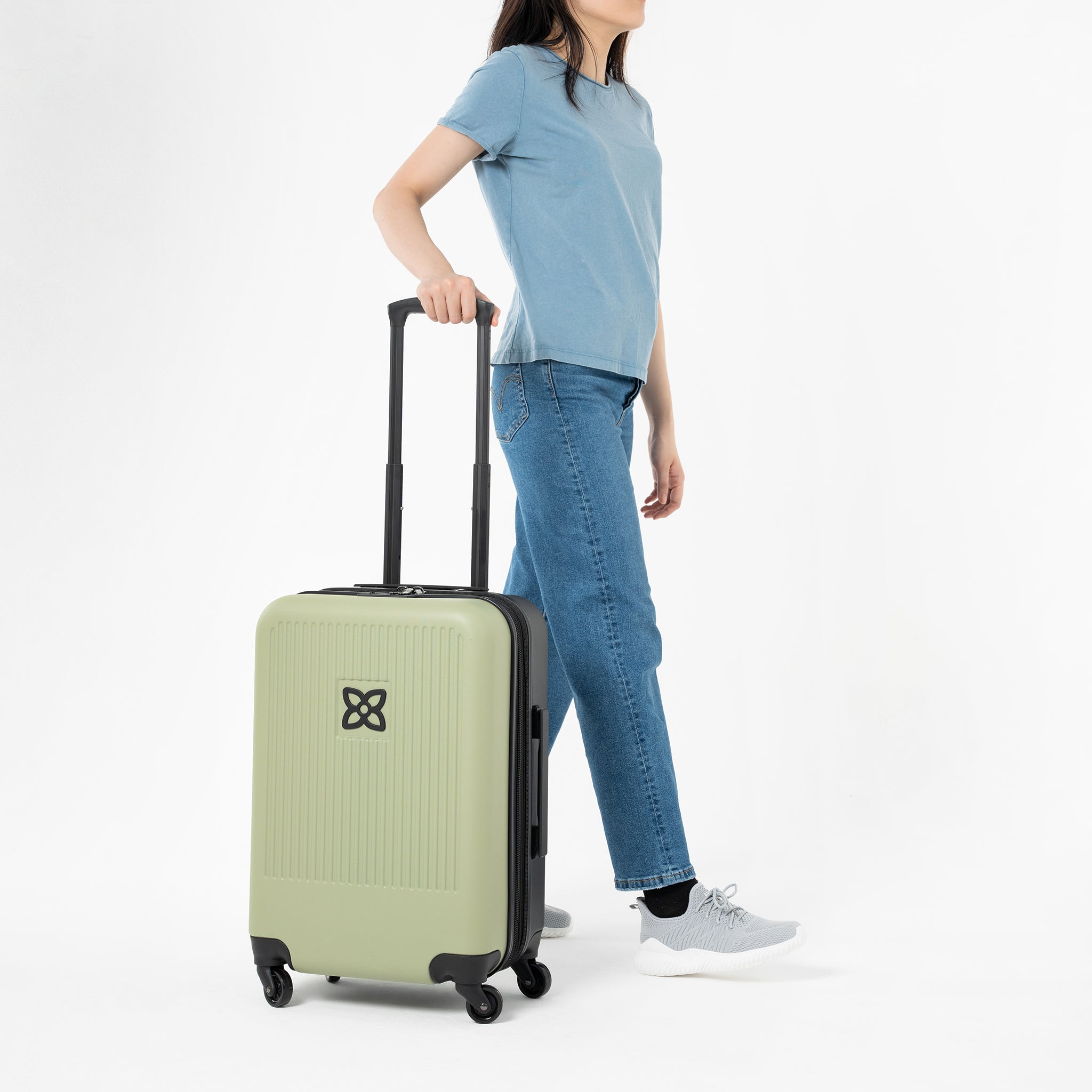 A model stands with one hand on the suitcase handle of Sherpani hard-shell carry-on suitcase, the Meridian in Sage. 