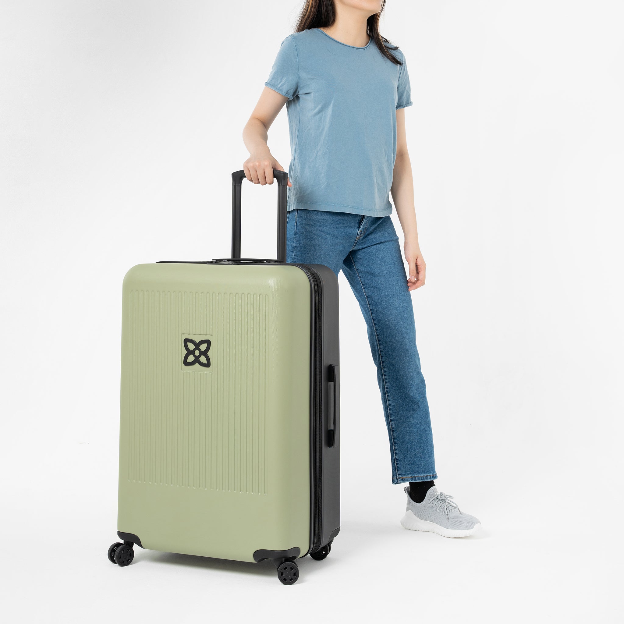 A model stands with one hand on the suitcase handle of Sherpani hard-shell luggage, the Meridian in Sage. #color_sage