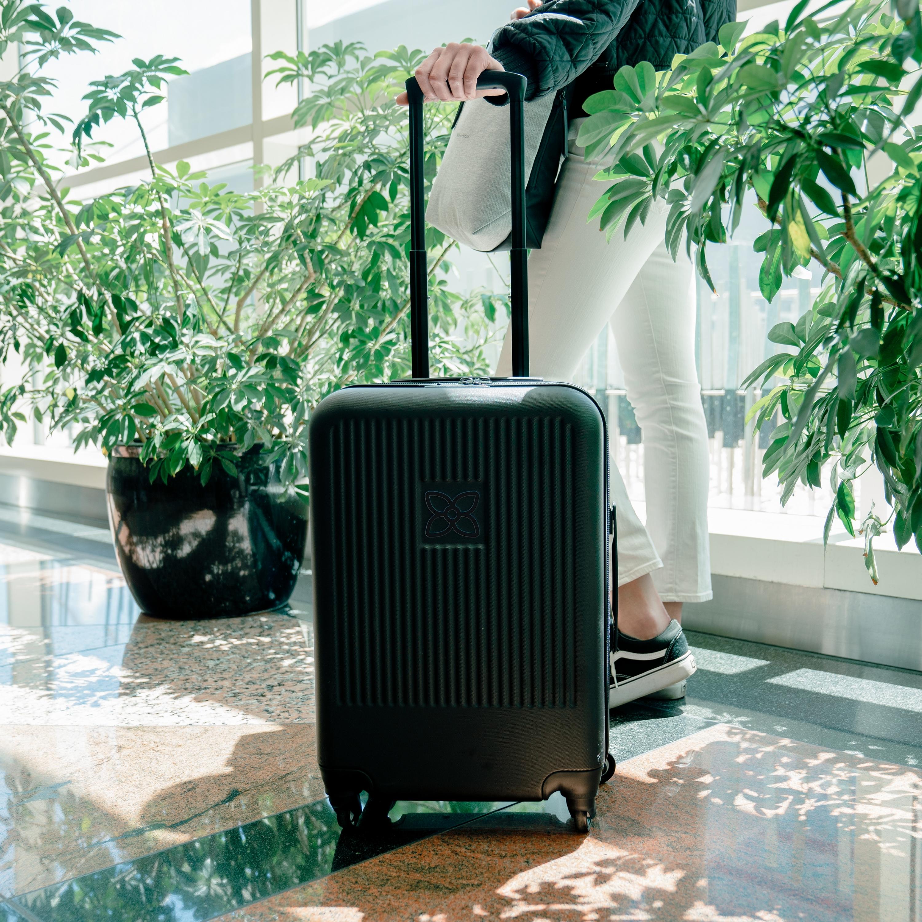 A woman waits at the airport with Sherpani hard shell luggage the Meridian. Part of Sherpani Travel Carry on Bundle in Classic Black. 