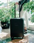 A woman waits at the airport with Sherpani hard-shell luggage the Meridian. Part of Sherpani Travel Carry-on Bundle in Classic Black.