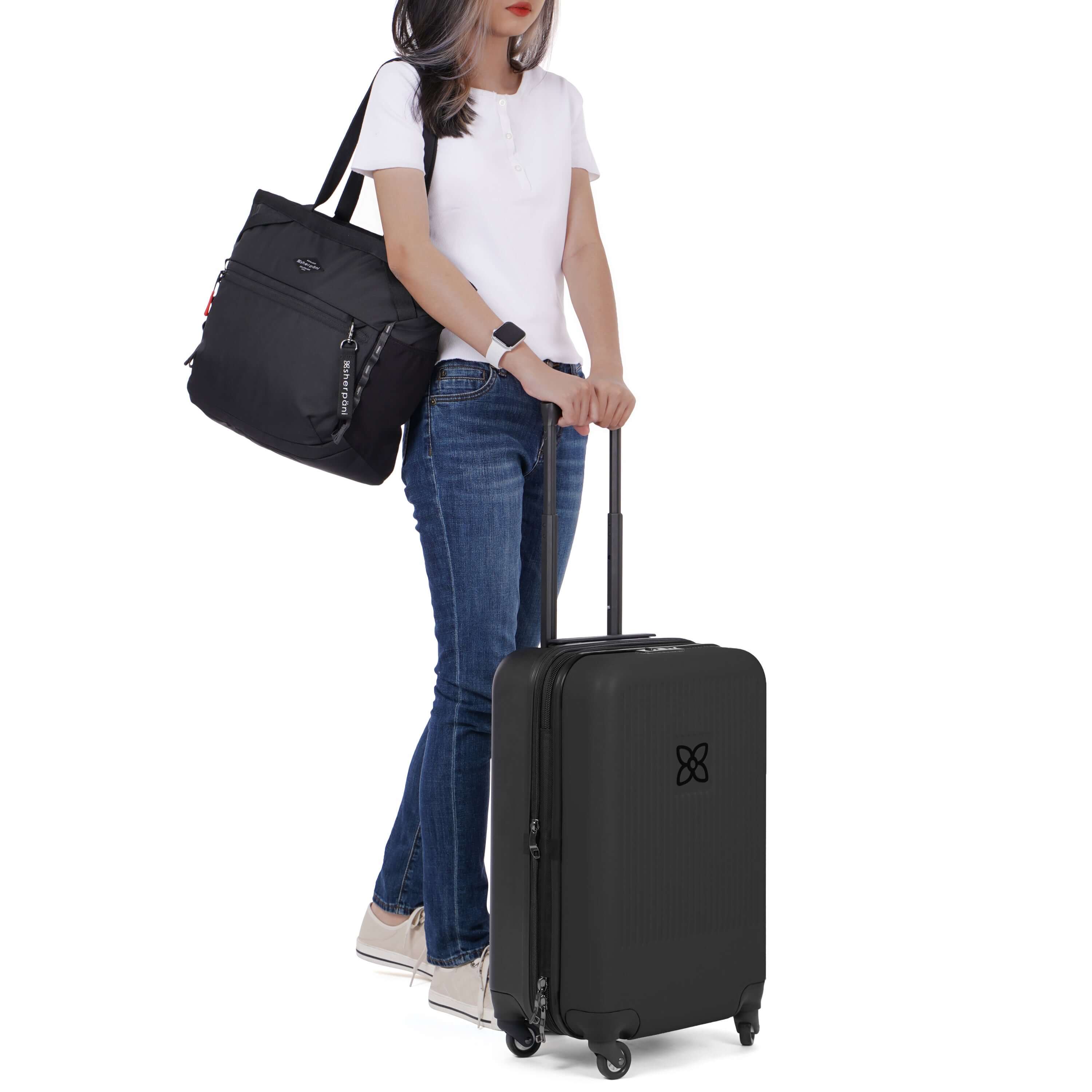A model stands with both hands on the retractable luggage handle of Sherpani suitcase, the Meridian in Black. Over her shoulder is Sherpani travel bag with trolley sleeve, the Stride in Raven. 