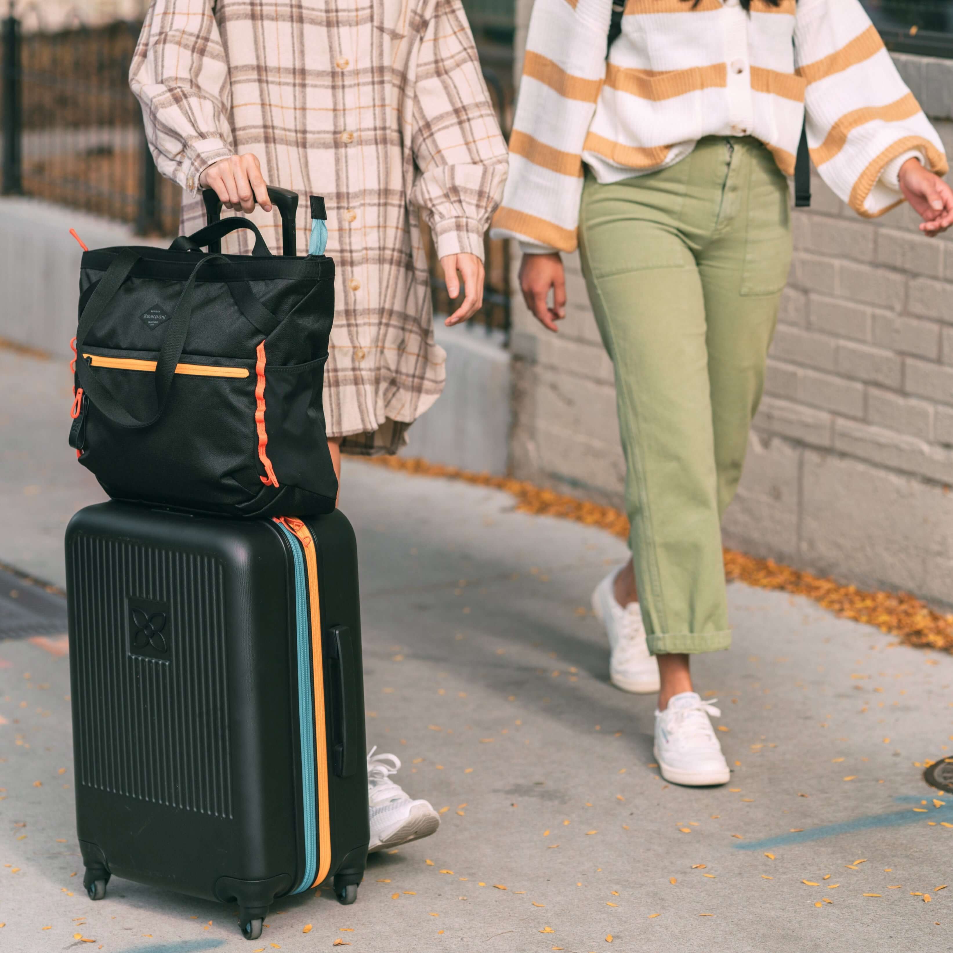Two women walking side-by-side. The woman on the left is pulling Sherpani hard-shell carry-on luggage, the Meridian in Chromatic. Atop the Meridian is Sherpani travel tote, the Stride in Chromatic, utilizing the luggage pass through feature. 