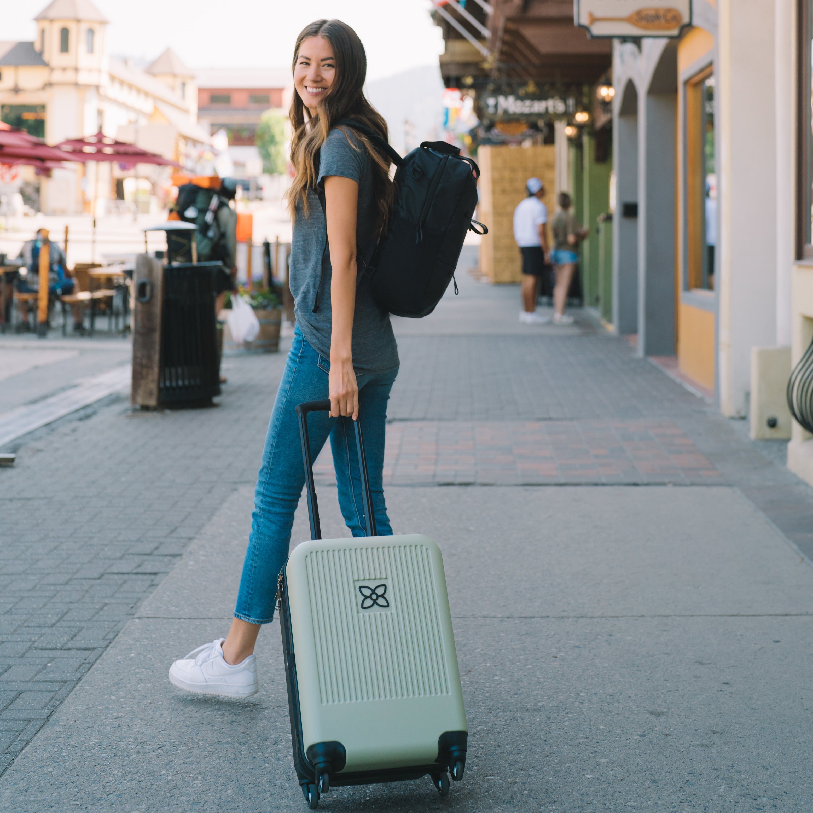 A woman standing on the street smiles at the camera. She is pulling Sherpani hard-shell carry-on suitcase, the Meridian in Sage, behind her. She is wearing Sherpani backpack with trolley sleeve, the Camden in Raven. 