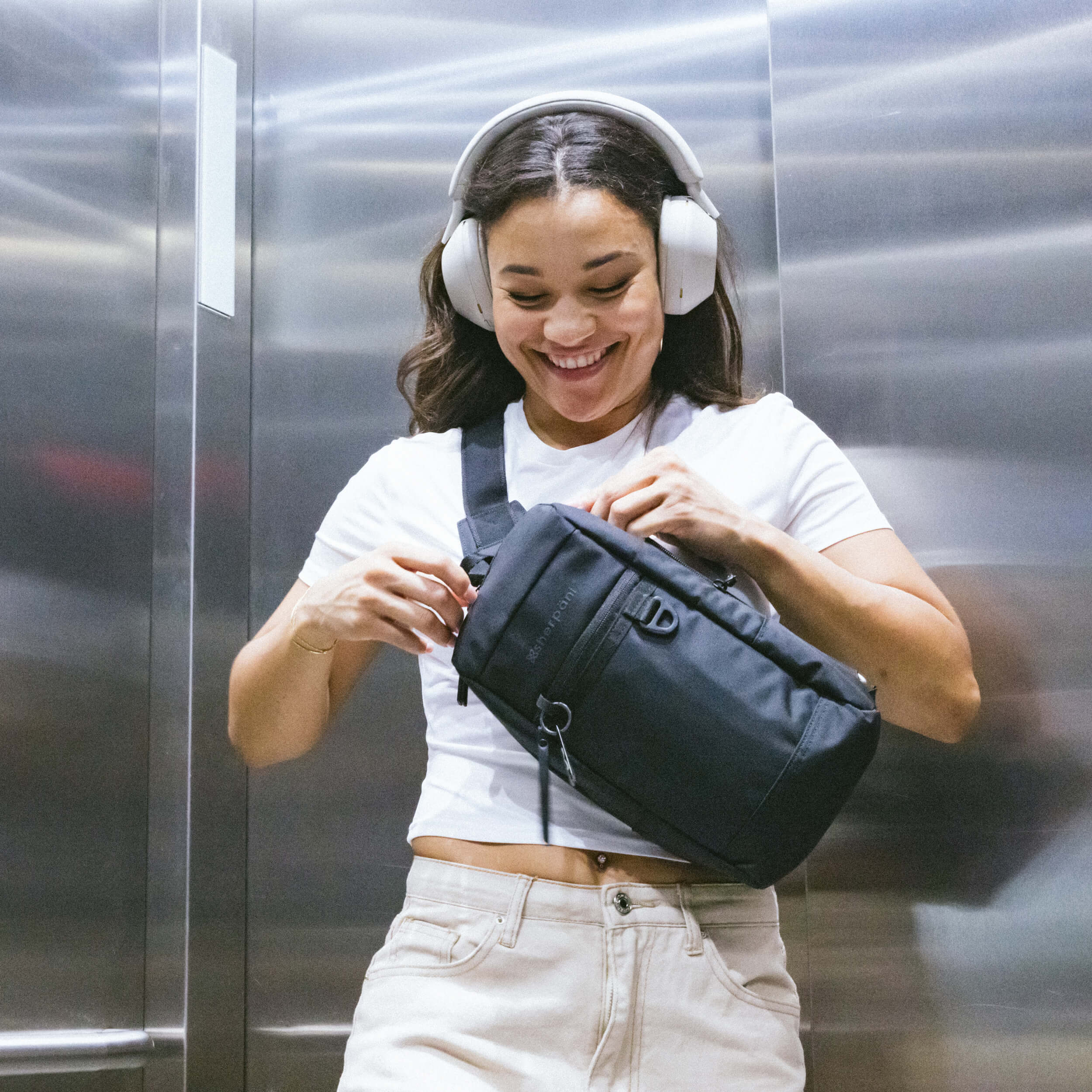 A woman in an elevator is unzipping her Anti-Theft sling bag, the Metro in Carbon. 