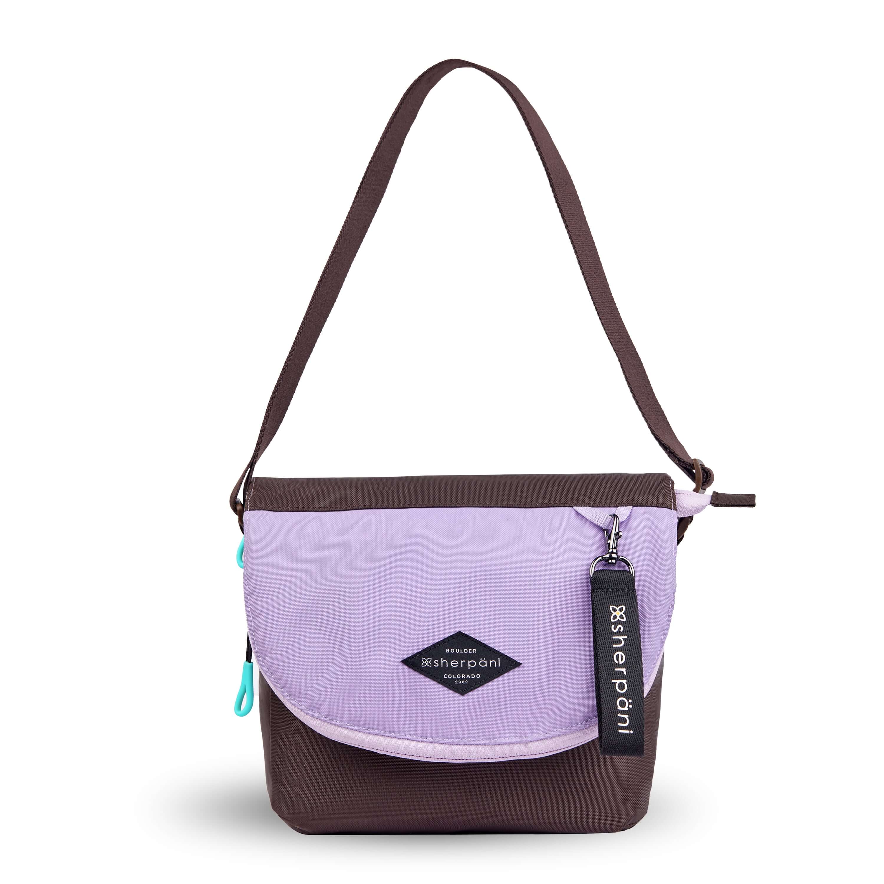 Flat front view of Sherpani crossbody, the Milli in Lavender. The messenger style bag is two-toned: the layover flap is lavender, the remainder of the bag is brown. Easy-pull zippers are accented in aqua. The bag has an adjustable crossbody strap. A branded Sherpani keychain is clipped to a fabric loop in the upper right corner.