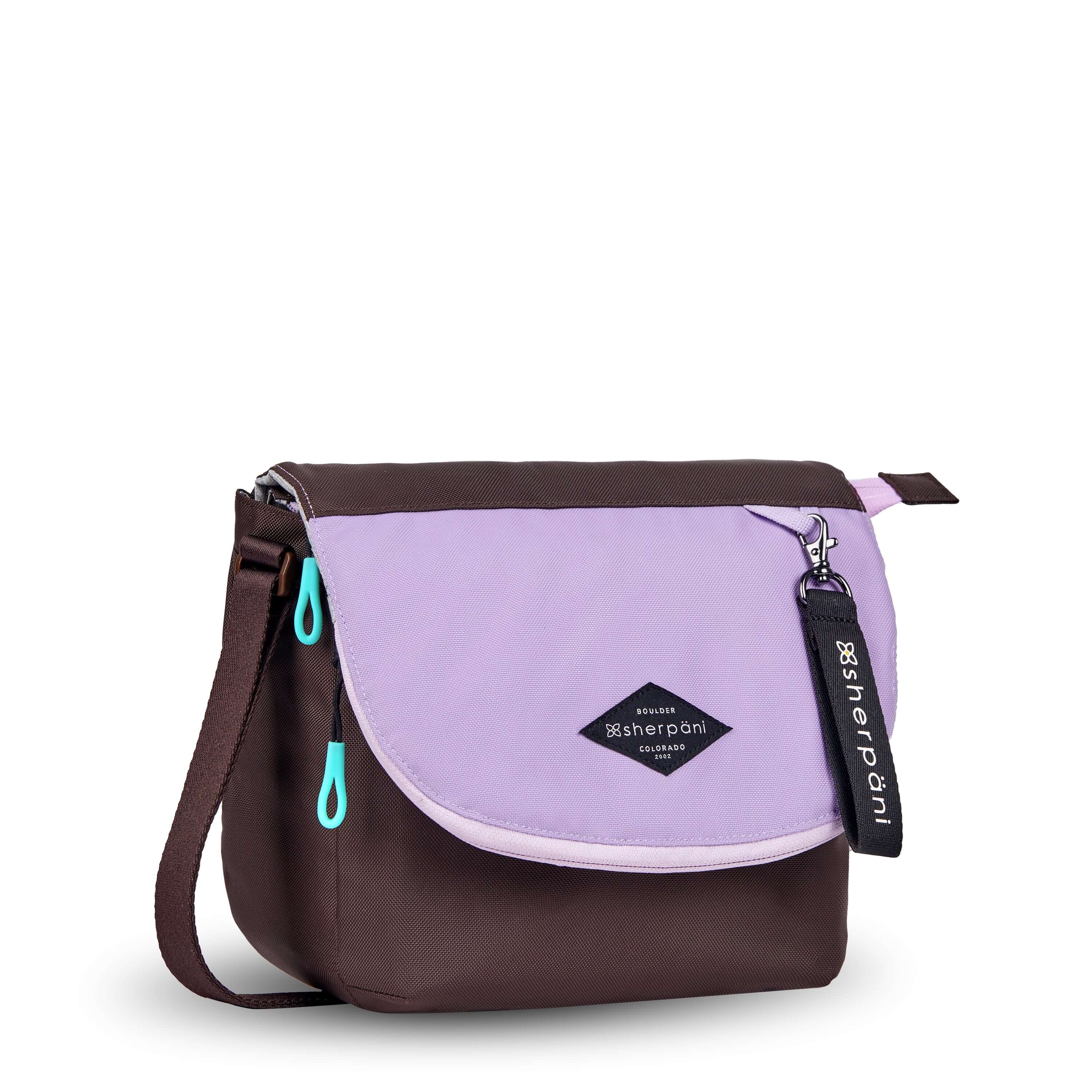 Angled front view of Sherpani crossbody, the Milli in Lavender. The messenger style bag is two-toned: the layover flap is lavender, the remainder of the bag is brown. Easy-pull zippers are accented in aqua. The bag has an adjustable crossbody strap. A branded Sherpani keychain is clipped to a fabric loop in the upper right corner.
