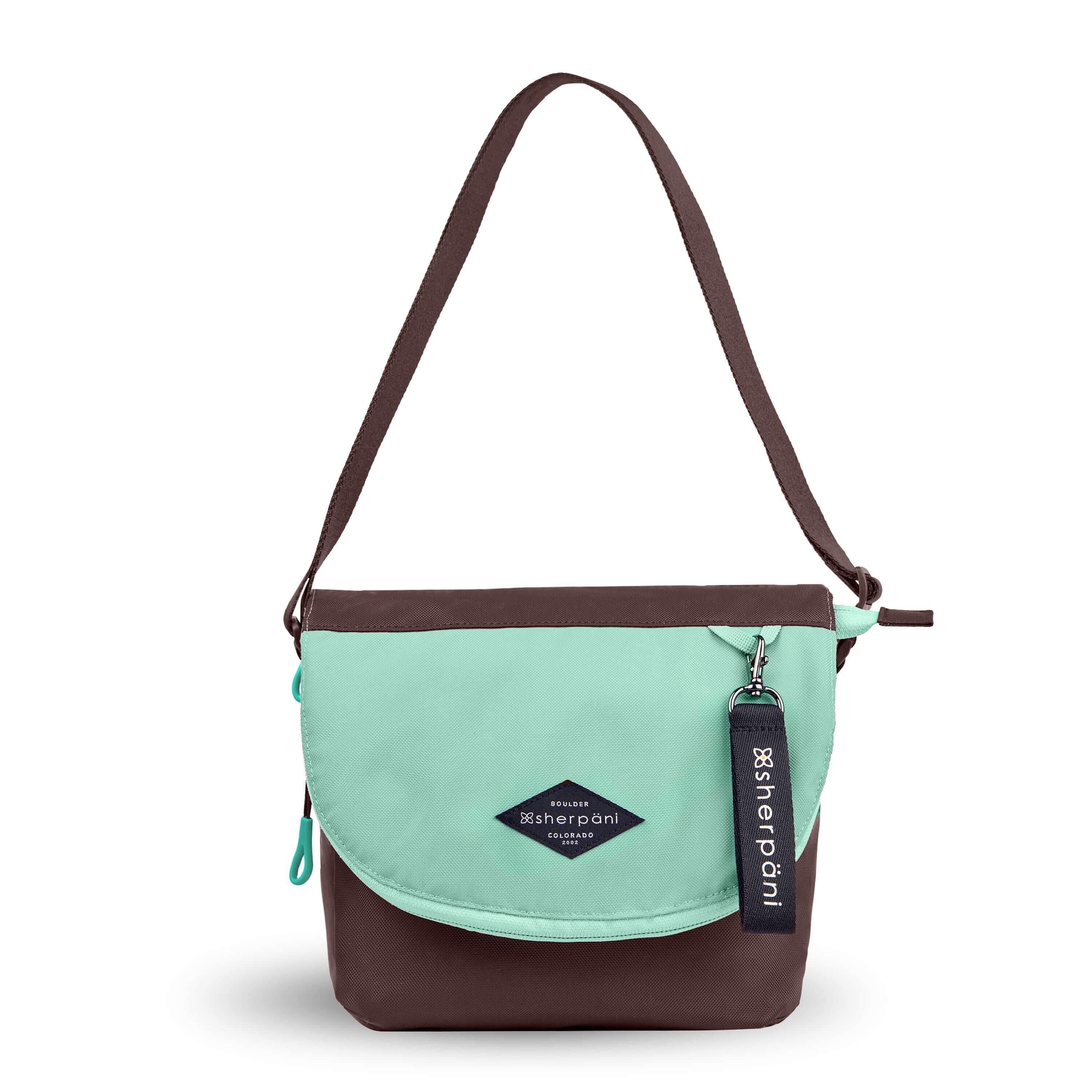 Flat front view of Sherpani crossbody, the Milli in Seagreen. The messenger style bag is two-toned: the layover flap is light green, the remainder of the bag is brown. Easy-pull zippers are accented in light green. The bag has an adjustable crossbody strap. A branded Sherpani keychain is clipped to a fabric loop in the upper right corner. 