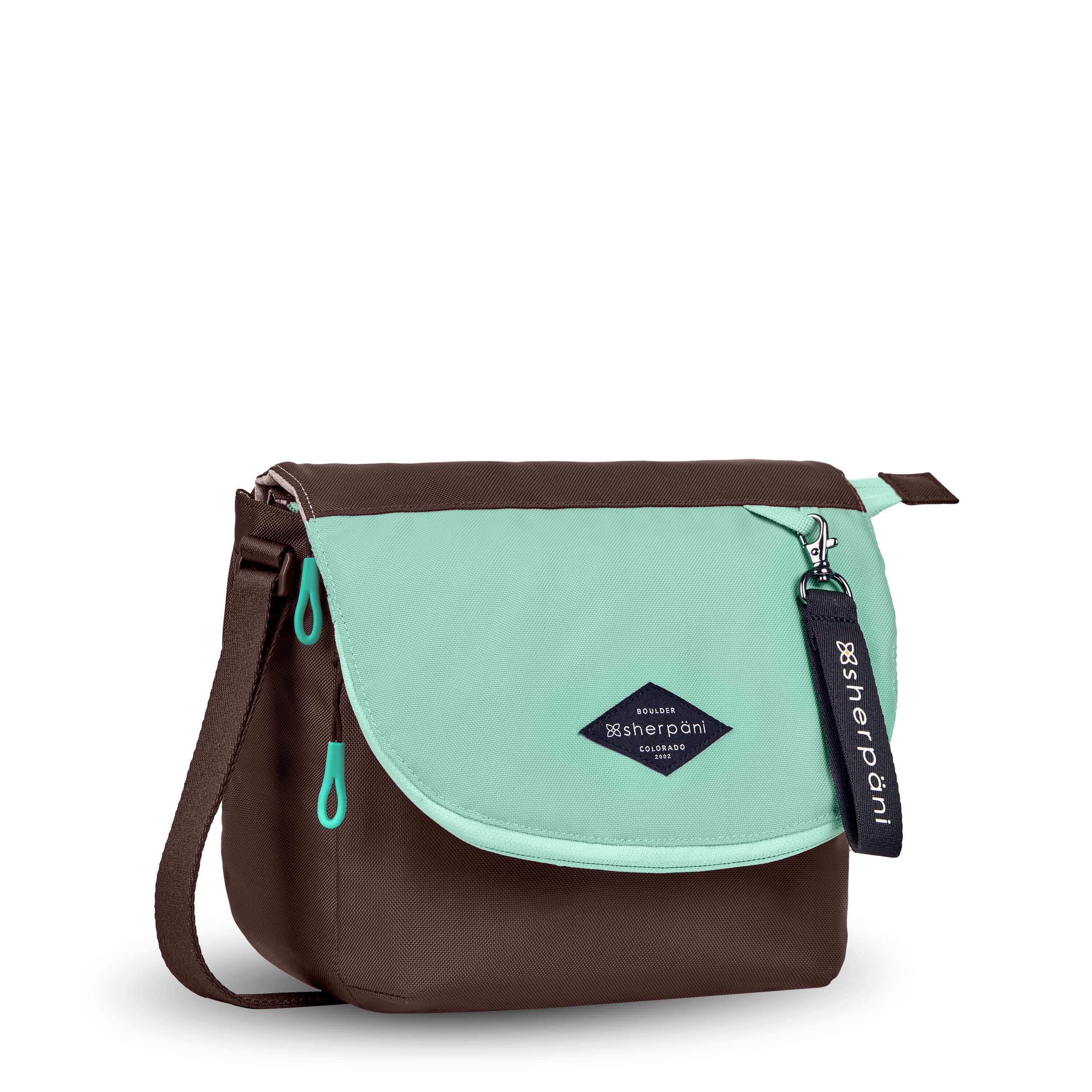 Angled front view of Sherpani crossbody, the Milli in Seagreen. The messenger style bag is two-toned: the layover flap is light green, the remainder of the bag is brown. Easy-pull zippers are accented in light green. The bag has an adjustable crossbody strap. A branded Sherpani keychain is clipped to a fabric loop in the upper right corner.