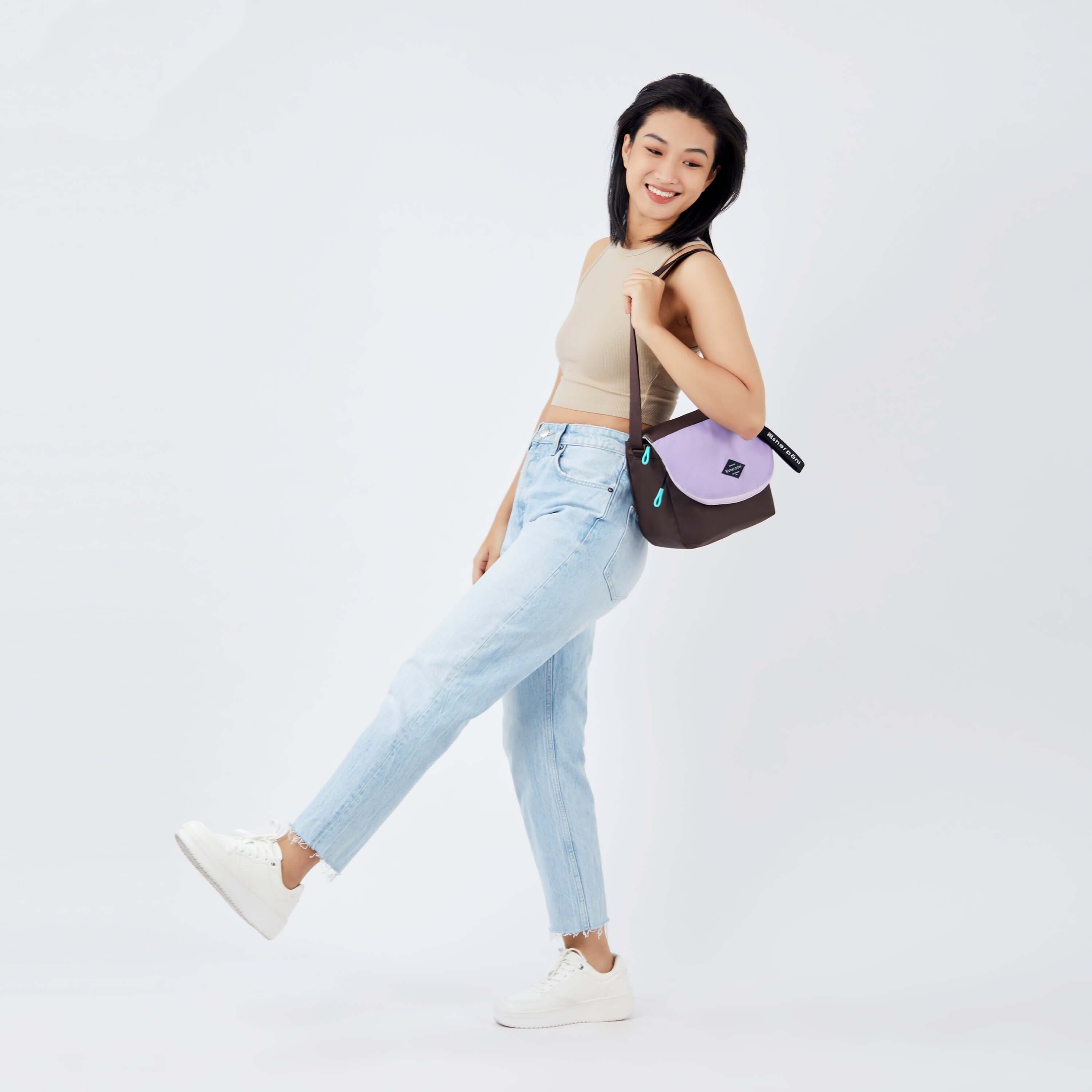 Full body view of a dark haired model facing the side and taking a dramatic step. She is wearing a beige crop top, jeans and white sneakers. She carries Sherpani crossbody, the Milli in Lavender, over her shoulder.