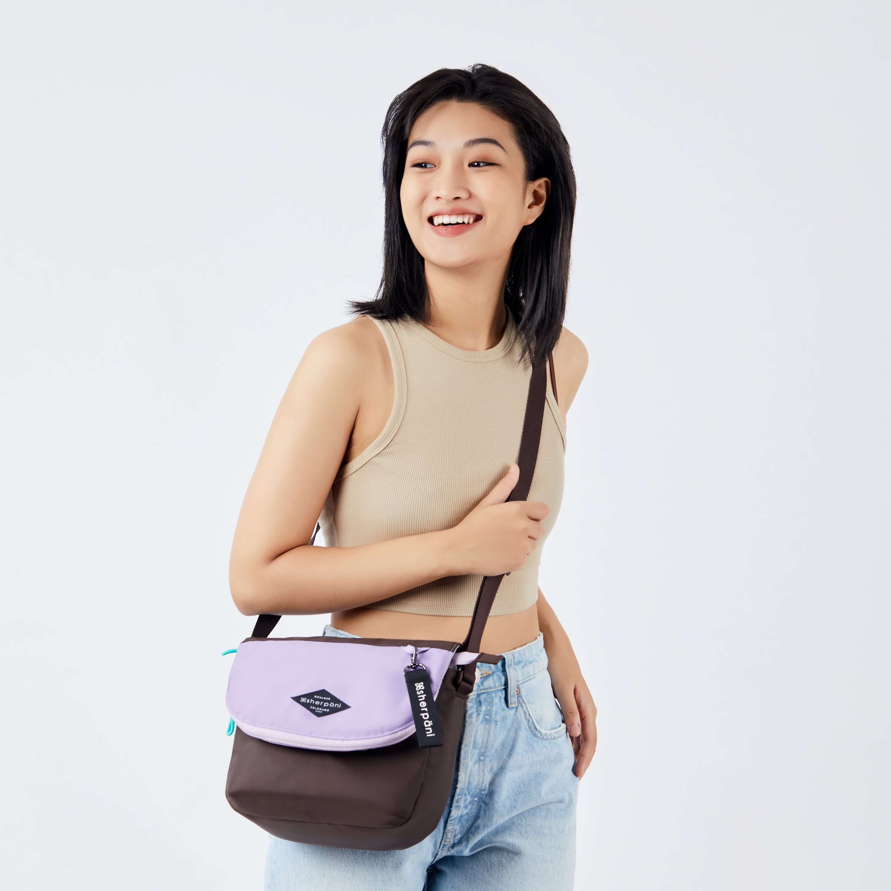 Close up view of a dark haired model facing the camera and smiling. She is wearing a beige crop top and jeans. She carries Sherpani crossbody, the Milli in Lavender, over her shoulder. #color_lavender