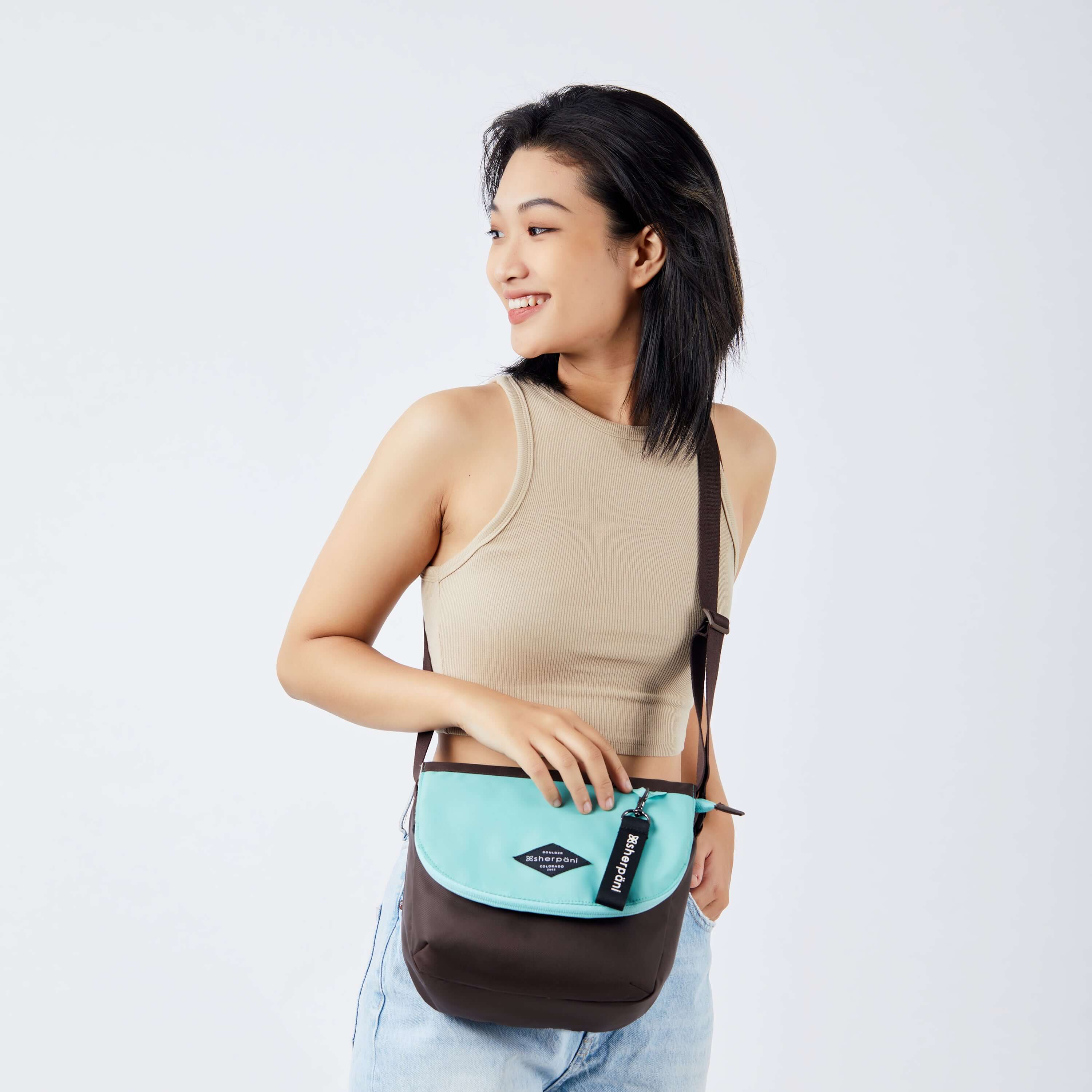 Close up view of a dark haired model facing the camera and smiling over her right shoulder. She is wearing a beige crop top and jeans. She carries Sherpani crossbody, the Milli in Seagreen, as a crossbody. #color_seagreen
