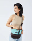 Close up view of a dark haired model facing the camera and smiling over her right shoulder. She is wearing a beige crop top and jeans. She carries Sherpani crossbody, the Milli in Seagreen, as a crossbody.