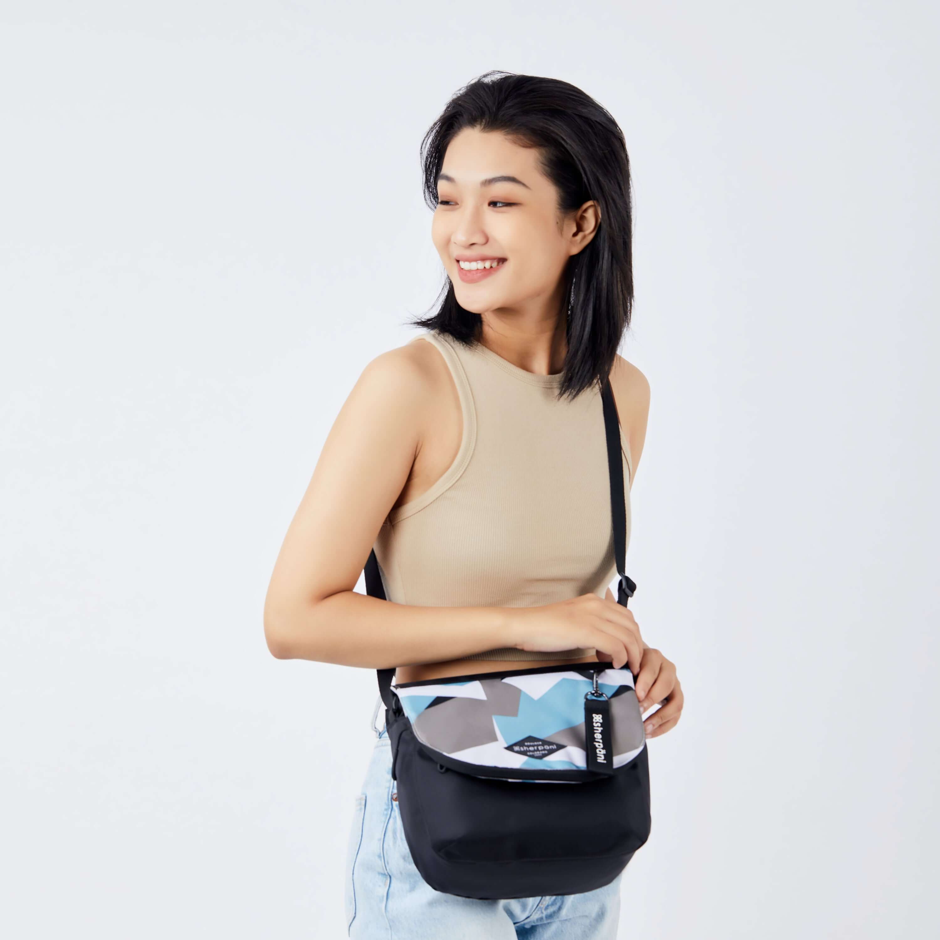 A dark haired model faces the camera and smiles over her right shoulder. She is wearing a beige crop top and jeans. She carries Sherpani crossbody, the Milli in Summer Camo, as a crossbody.