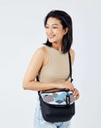 A dark haired model faces the camera and smiles over her right shoulder. She is wearing a beige crop top and jeans. She carries Sherpani crossbody, the Milli in Summer Camo, as a crossbody.