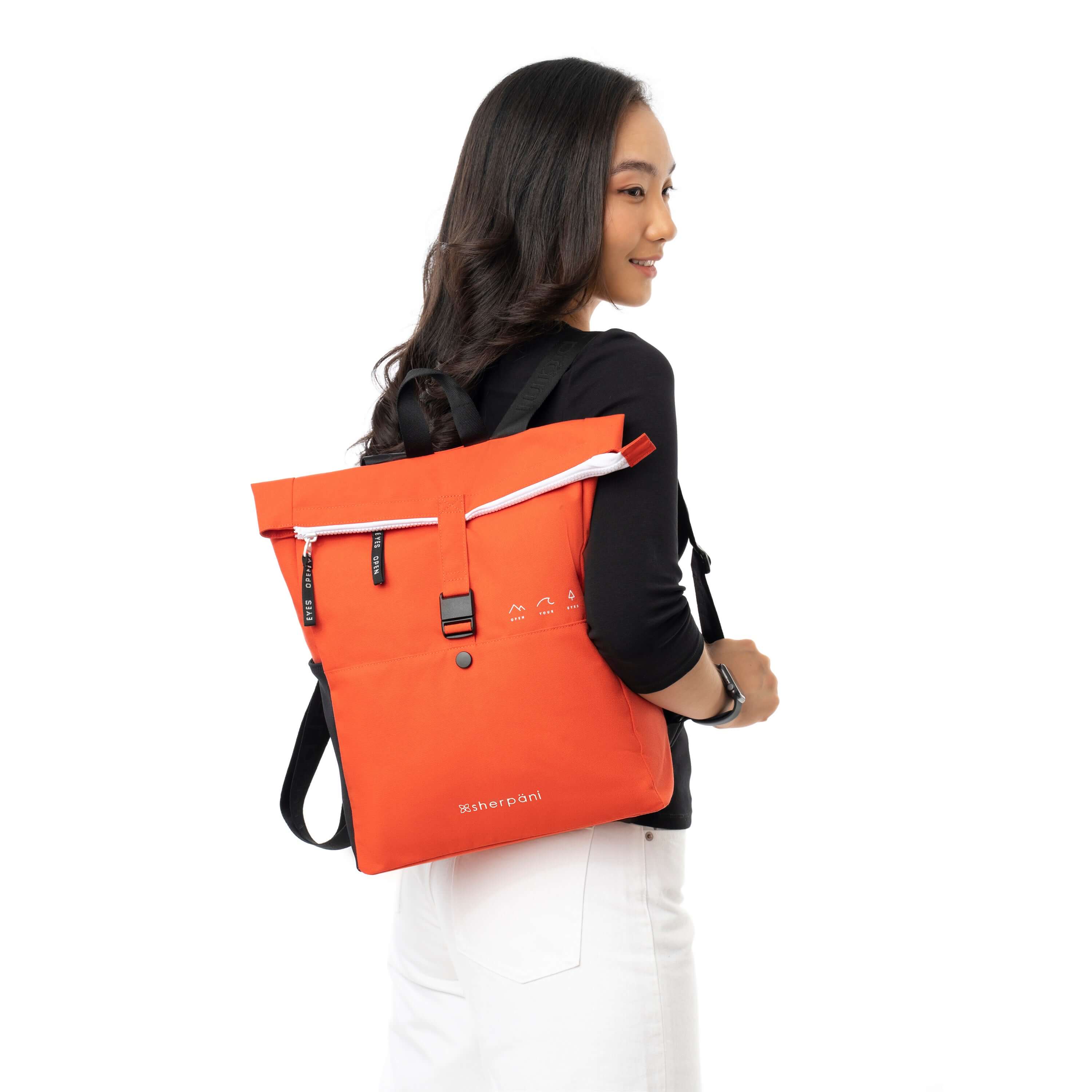 A dark haired model facing away from the camera and smiling over her right shoulder. She is wearing a black shirt and white pants. She carries Sherpani backpack, the Miyako in Poppy, over one shoulder.