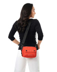 A dark haired model facing away from the camera and looks over her right shoulder. She is wearing a black shirt and white pants. She carries Sherpani crossbody, the Osaka in Poppy, over her shoulder.