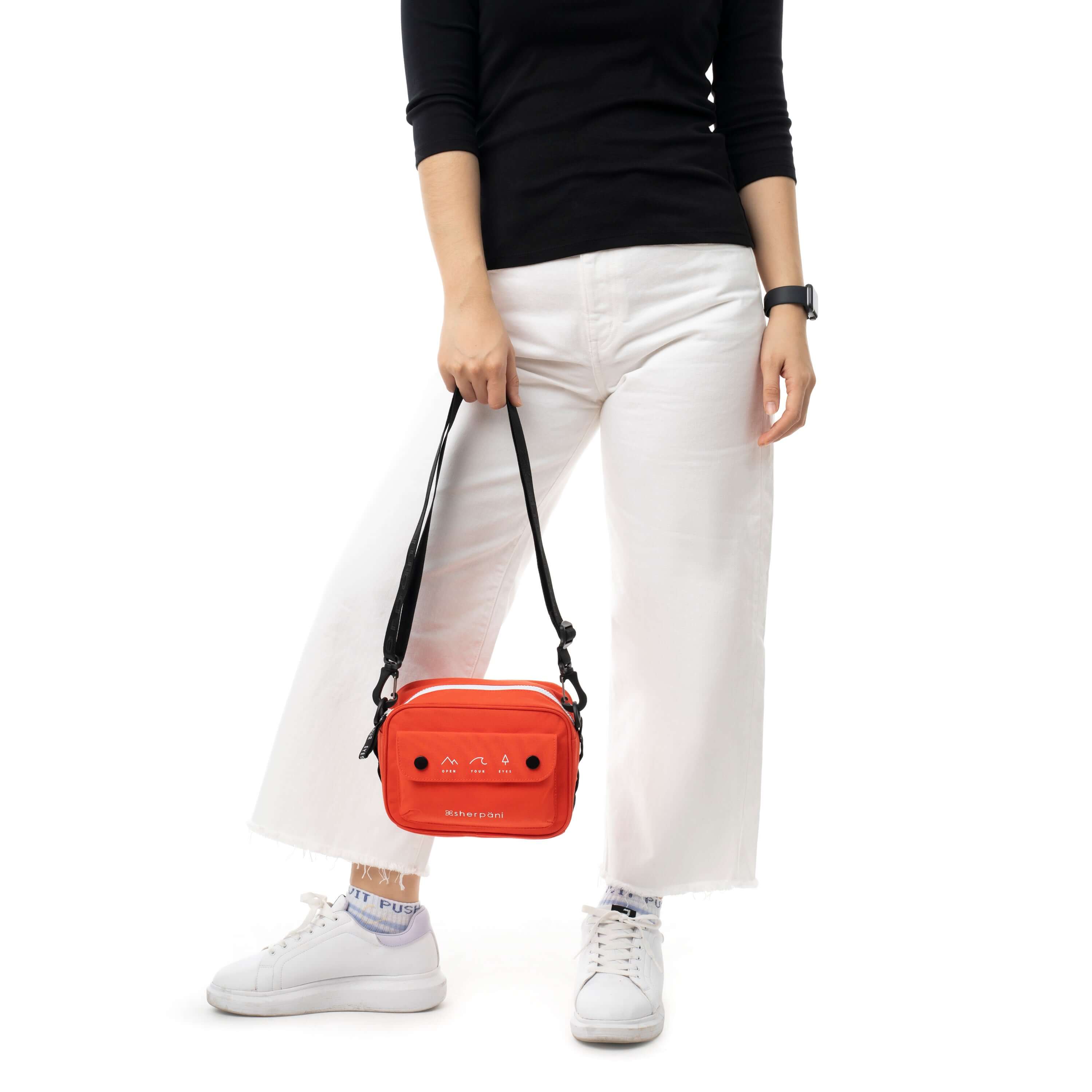 Close up view of a model facing the camera. She is wearing a black shirt, white pants and white sneakers. She carries Sherpani crossbody, the Osaka in Poppy, by the adjustable/detachable crossbody strap.