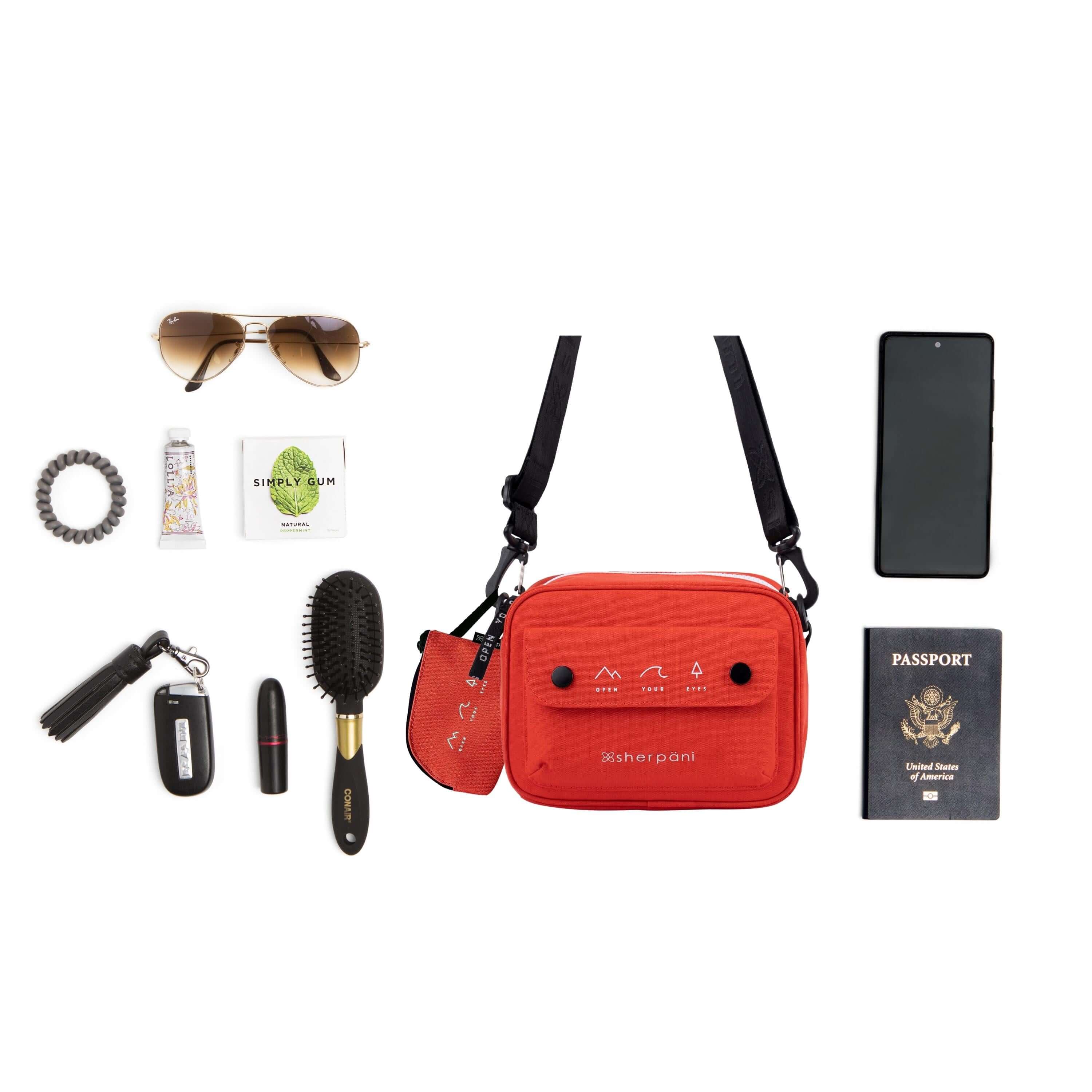 Top view of example items to fill the bag. Sherpani crossbody, the Osaka in Poppy, lies in the center. It is surrounded by an assortment of items: sunglasses, hair tie, hand lotion, gum, car key, lipstick, hairbrush, phone and passport.
