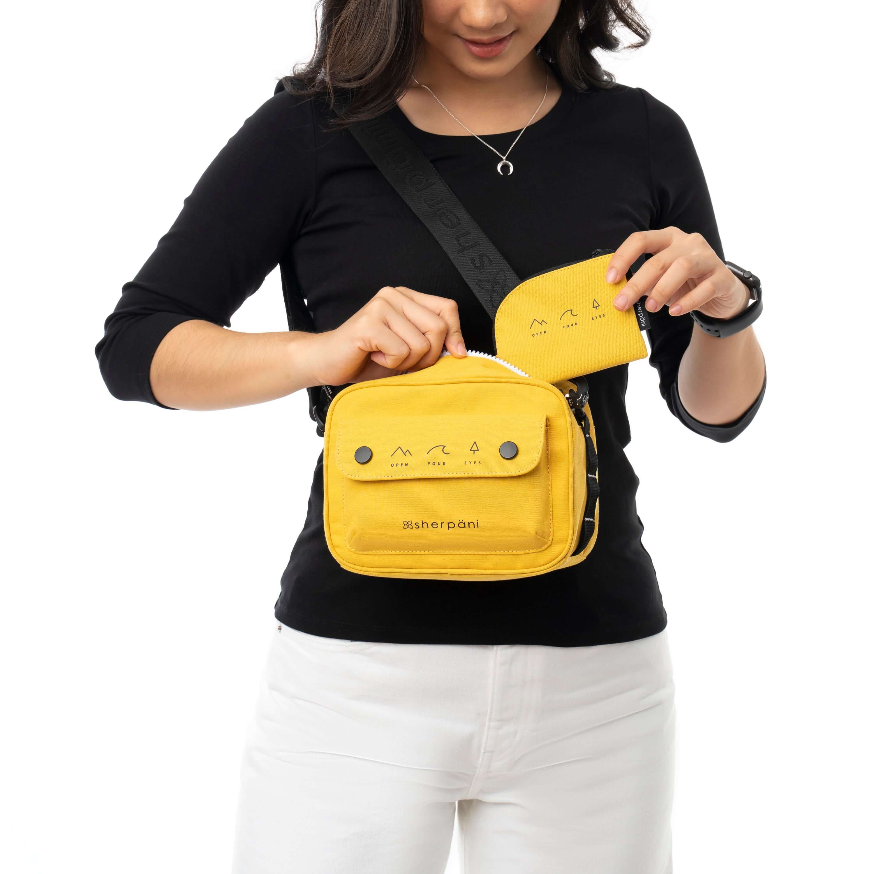 Close up view of a brown haired woman facing the camera and looking down at her bag. She is wearing a black shirt and white pants. She carries Sherpani crossbody, the Osaka in Sunflower, over her shoulder. She is pulling the detachable coin purse out of the bag.