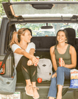Two women share snacks and smiles in the back of a parked car on a summer day. The woman on the left has blonde hair, she is wearing a white tee shirt and black leggings. Next to her, sits Sherpani mesh crossbody, the Payton. The woman on the right has brown hair, she is wearing a green tank top and jeans. In between the two women, sits Sherpani mesh backpack, the Adaline.