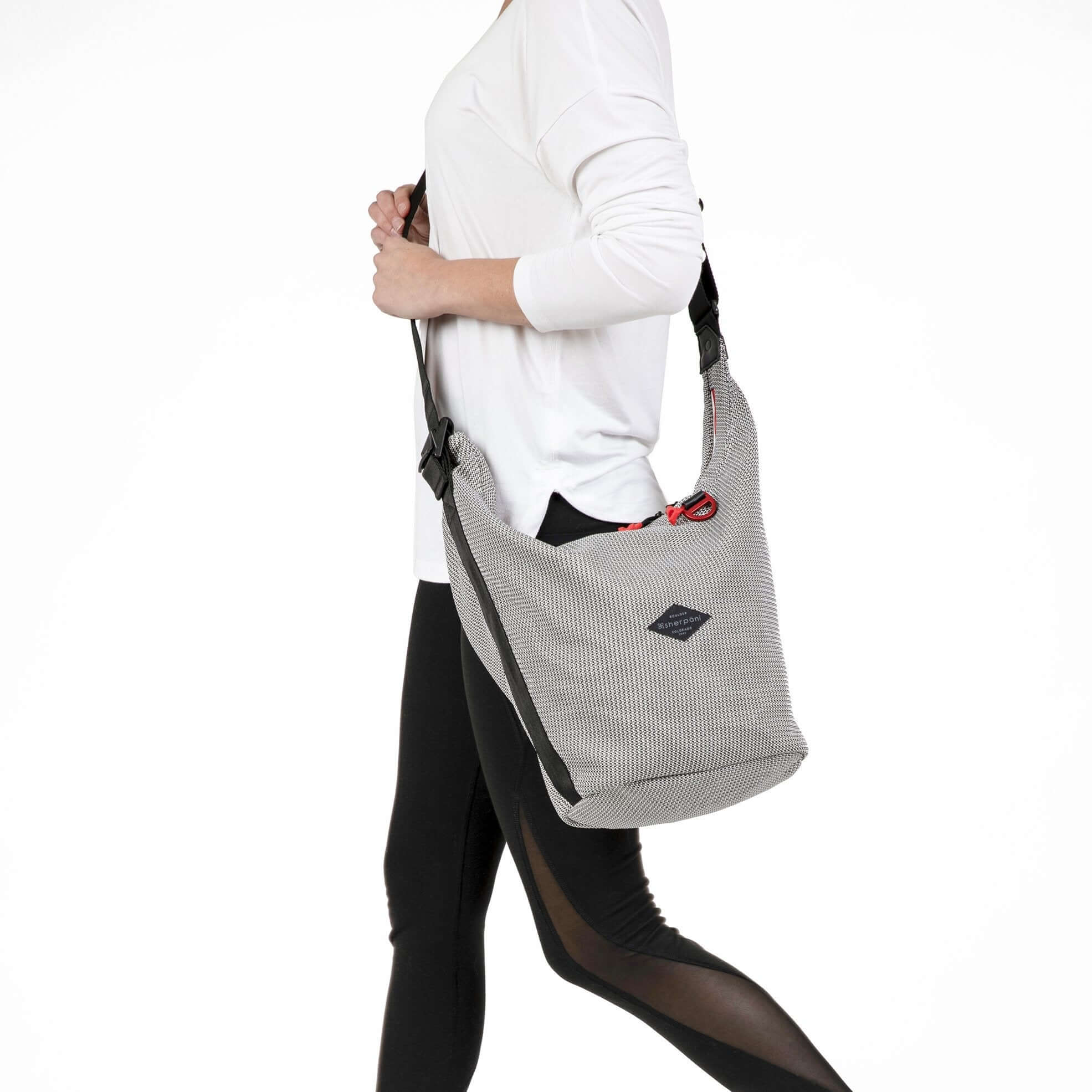 Close up view of a model facing the side and walking. She is wearing a long sleeved white shirt and black leggings. She carries Sherpani mesh crossbody, the Payton.