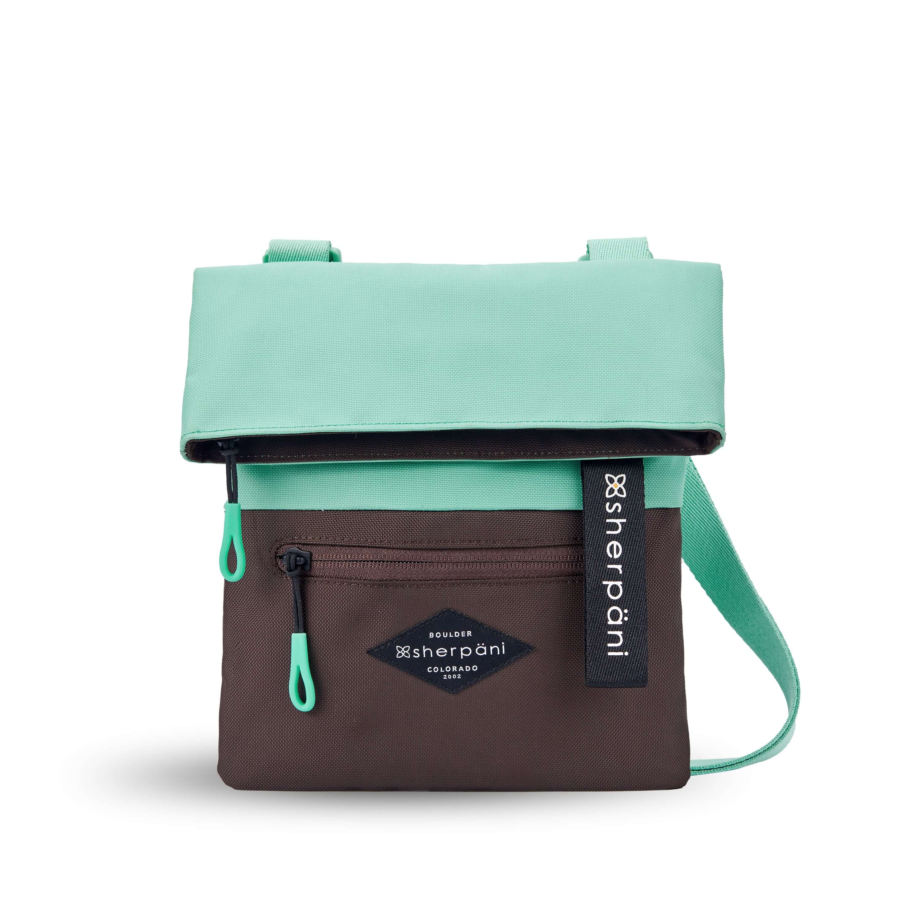 Flat front view of Sherpani crossbody, the Pica in Seagreen. The bag is two toned: the top half is light green and the bottom half is brown. There is an external zipper pocket on the front panel. Easy-pull zippers are accented in light green. The top of the bag folds over creating a signature look. A branded Sherpani keychain is clipped to the upper right corner. The bag has an adjustable crossbody strap. 