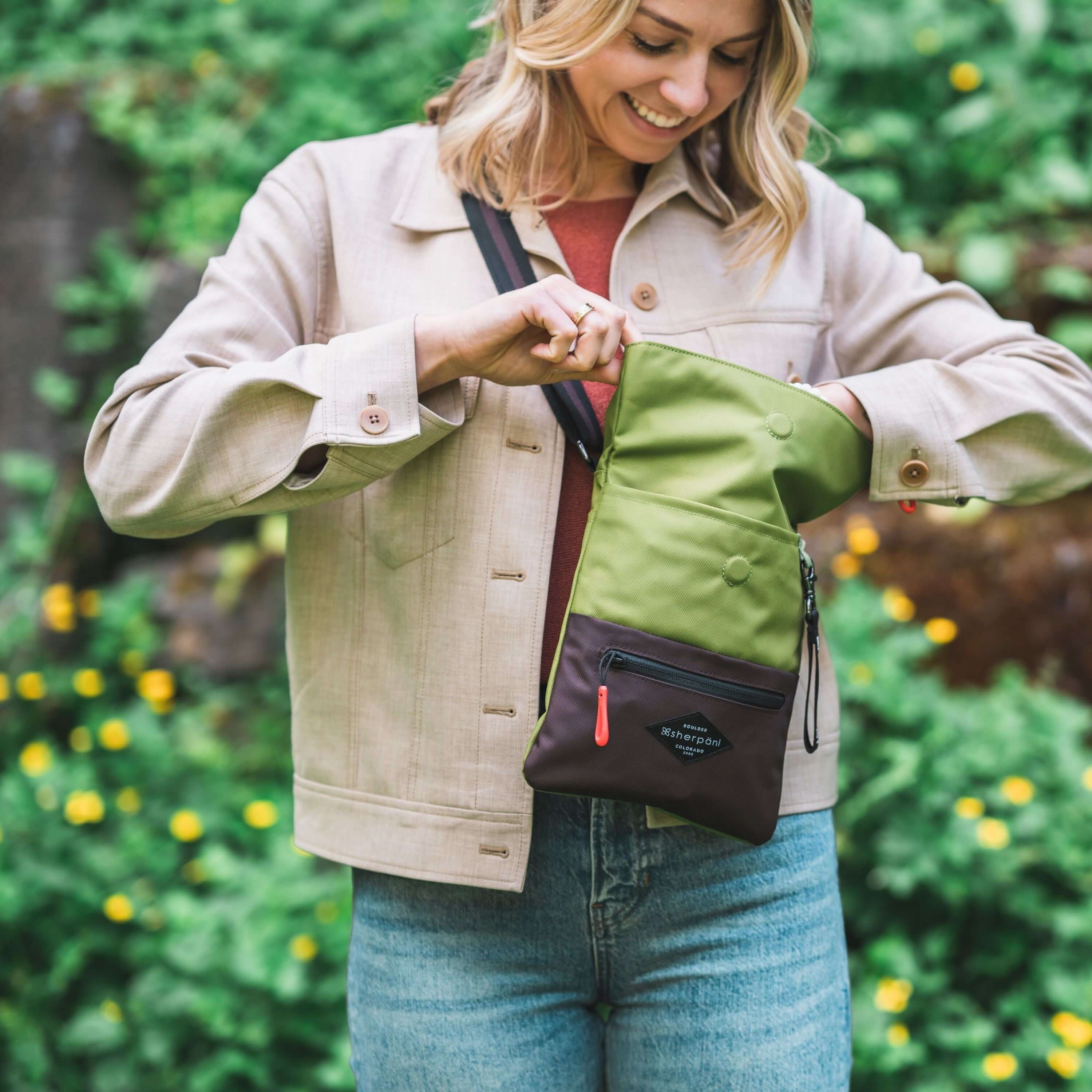 A blonde woman stands outside in front of a bush with yellow flowers. She is wearing a tan jacket and jeans. She is smiling down into the main compartment of her Sherpani crossbody, the Pica in Cactus. 