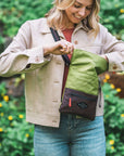 A blonde woman stands outside in front of a bush with yellow flowers. She is wearing a tan jacket and jeans. She is smiling down into the main compartment of her Sherpani crossbody, the Pica in Cactus.