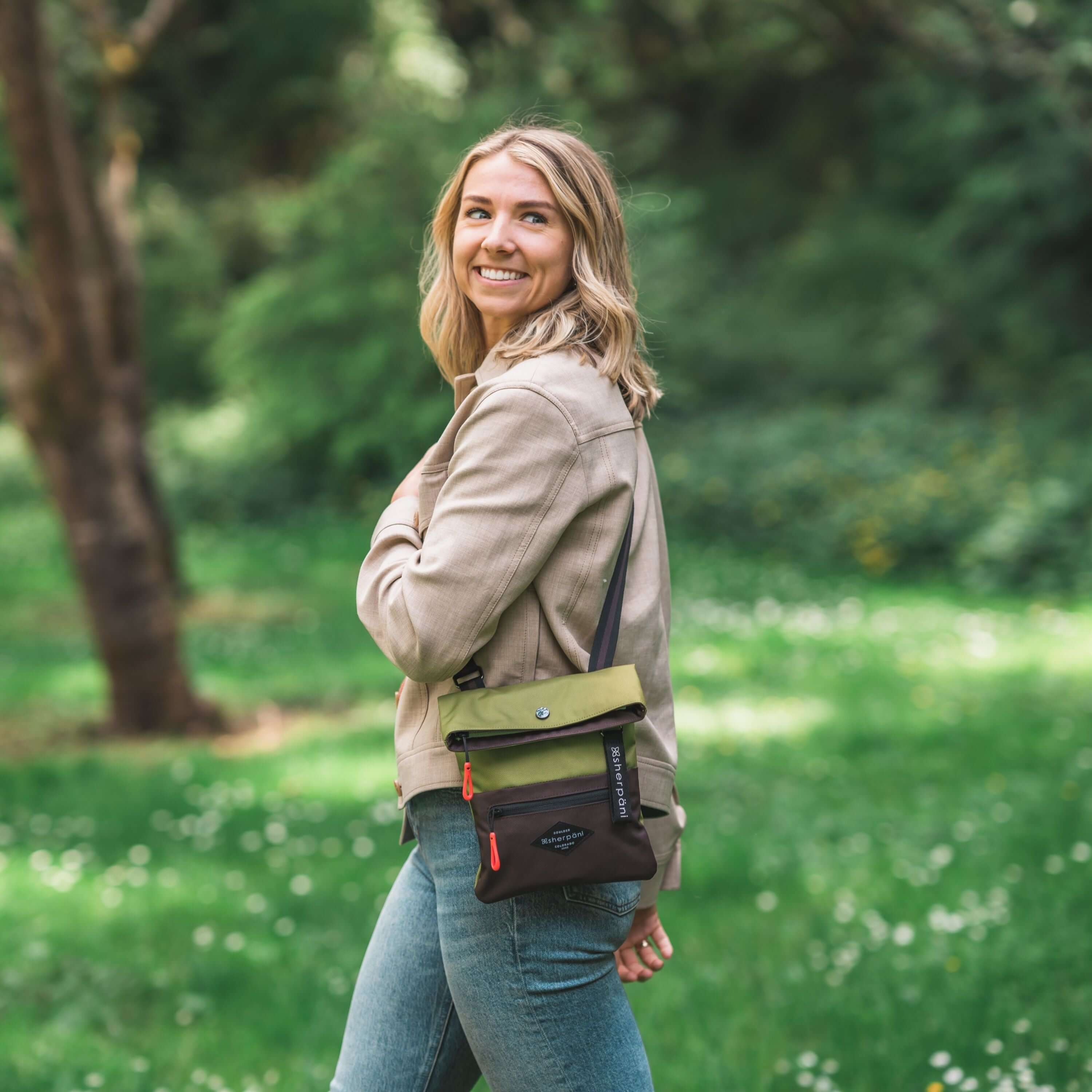 A blonde woman smiles while walking outside in a park. She is wearing a tan jacket and jeans. She carries Sherpani crossbody, the Pica in Cactus. 