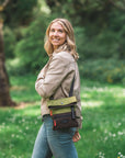 A blonde woman smiles while walking outside in a park. She is wearing a tan jacket and jeans. She carries Sherpani crossbody, the Pica in Cactus.