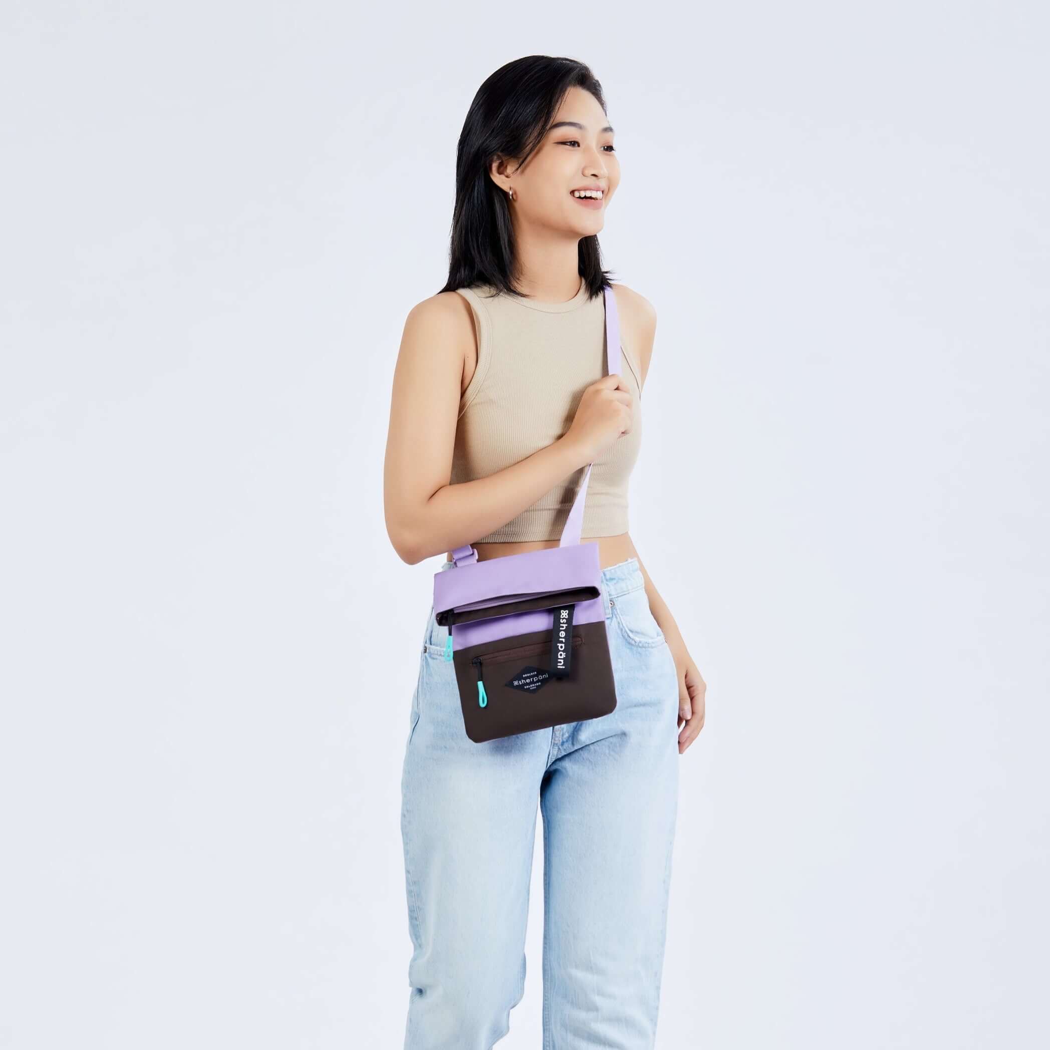 Full body view of a dark haired model facing the camera and smiling to the side. She is wearing a tan crop top, jeans and white sneakers. She carries Sherpani crossbody, the Pica in lavender, as a crossbody.