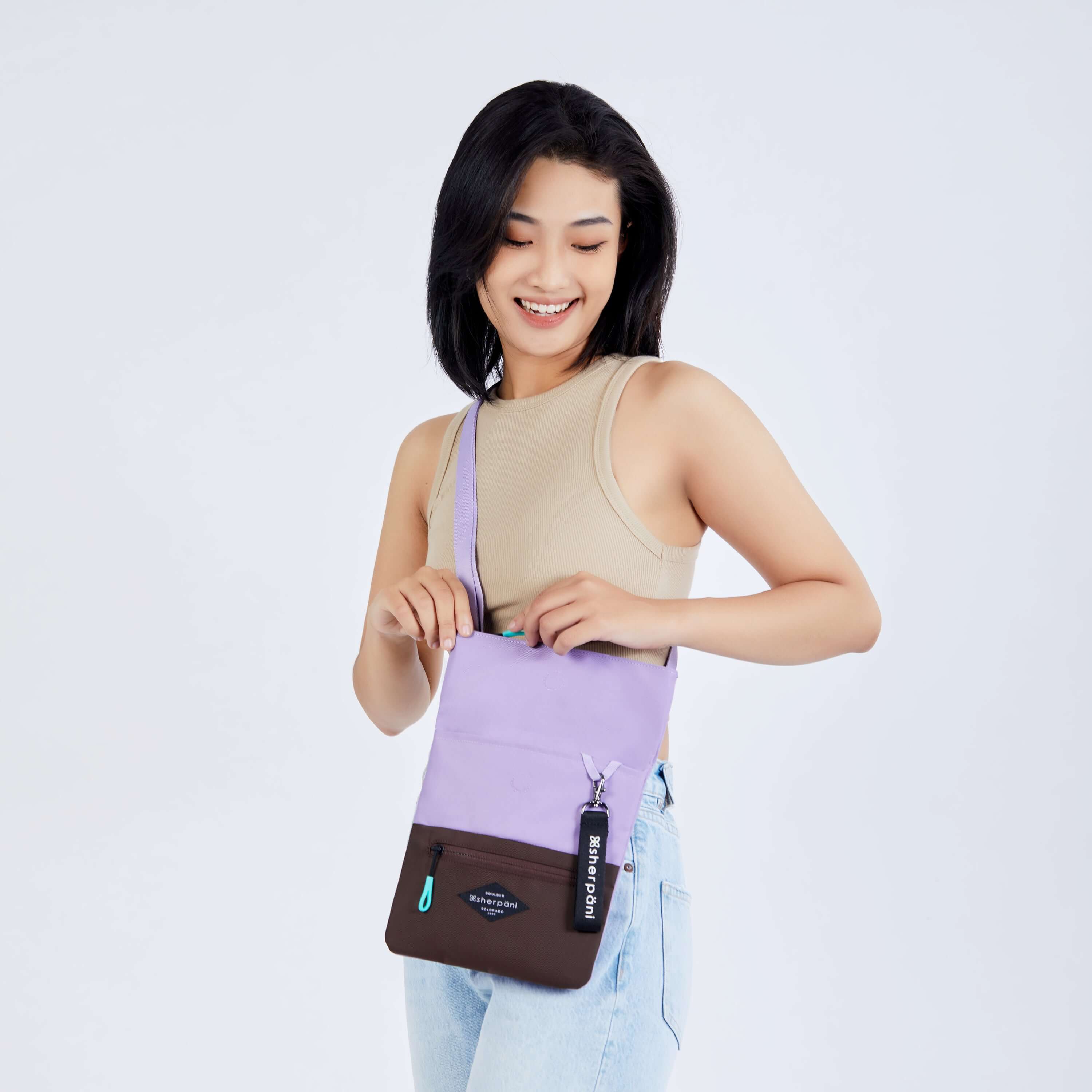 Close up view of a dark haired model facing the camera and smiling downward. She is wearing a tan crop top and jeans. She is unzipping the main compartment of Sherpani crossbody, the Pica in Lavender. 