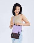 Close up view of a dark haired model facing the camera and smiling downward. She is wearing a tan crop top and jeans. She is unzipping the main compartment of Sherpani crossbody, the Pica in Lavender.