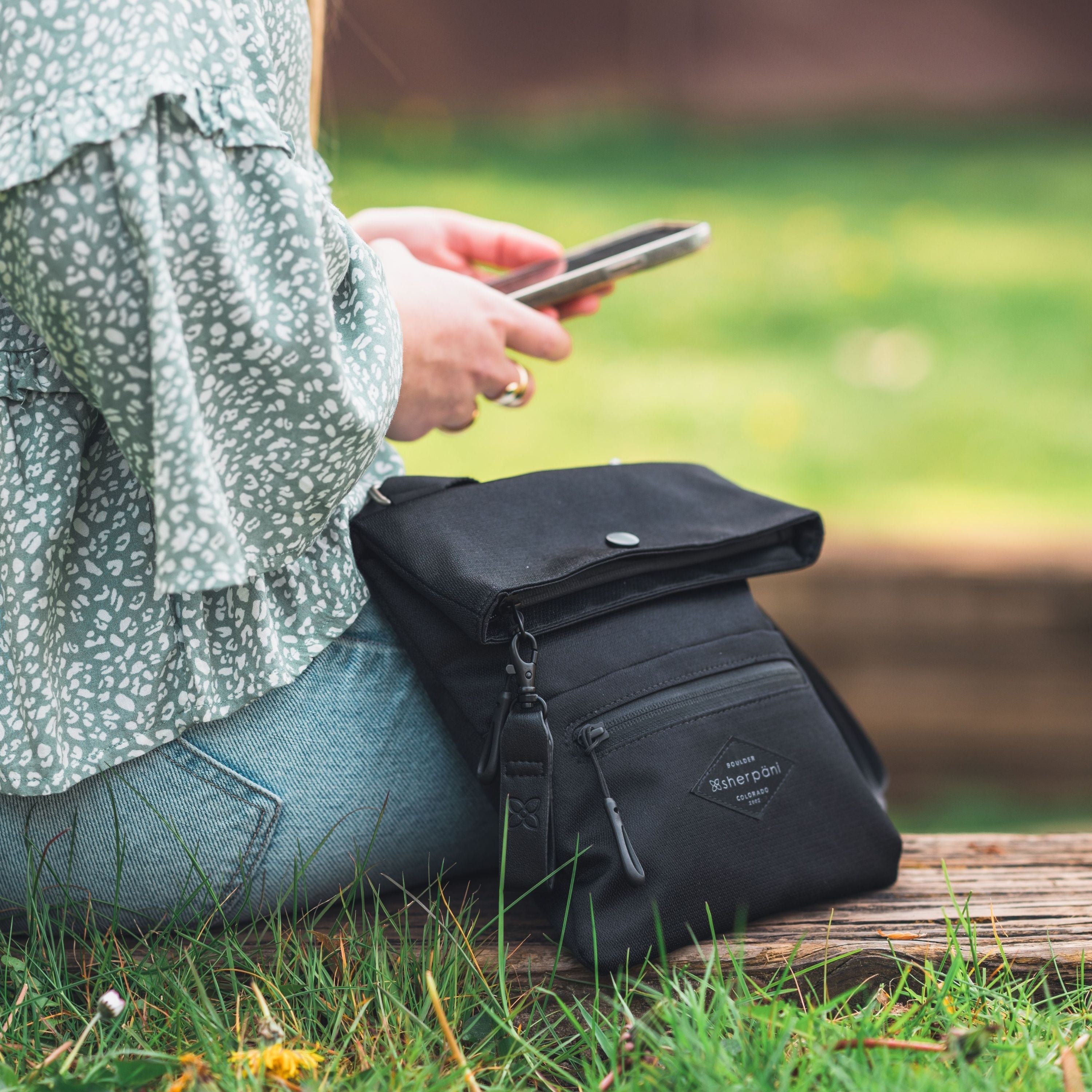 Close up view of a woman sitting on the grass outside. She is wearing a green top and jeans. She is playing on her phone. Next to her sits Sherpani crossbody, the Pica in Raven.