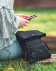 Close up view of a woman sitting on the grass outside. She is wearing a green top and jeans. She is playing on her phone. Next to her sits Sherpani crossbody, the Pica in Raven.