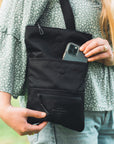 Close up view of a blonde woman outside. She is wearing a green top and jeans. She is putting her phone into the external pouch on the front of Sherpani crossbody, the Pica in Raven.