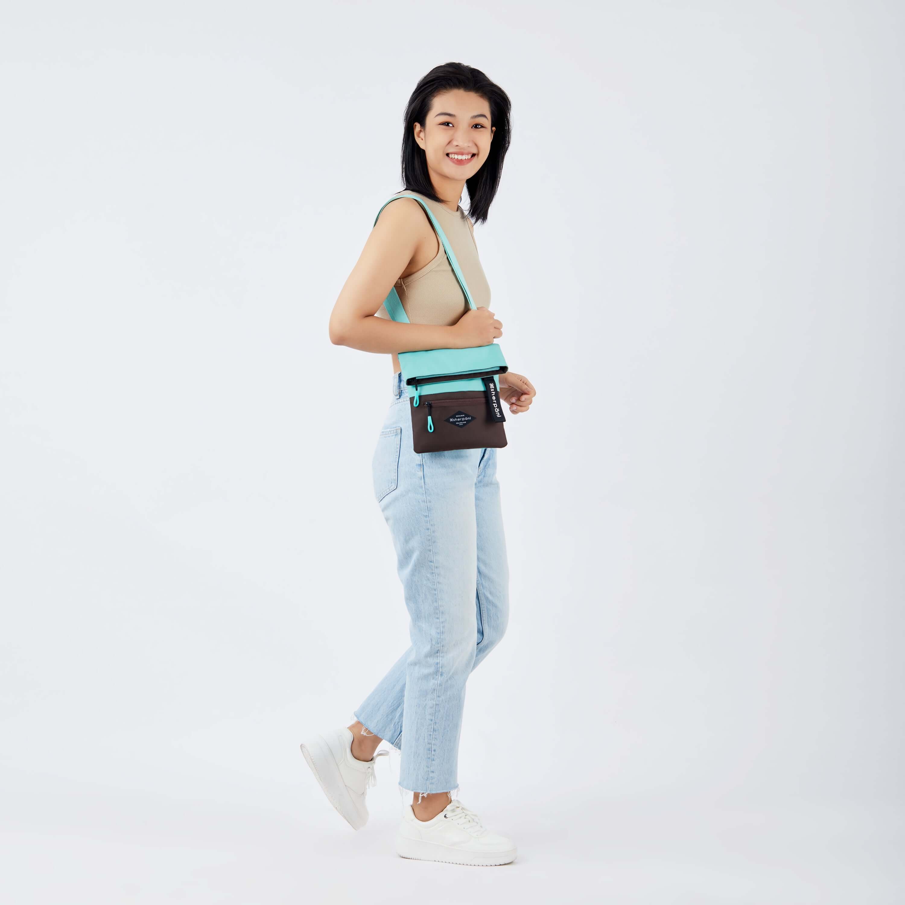 Full body view of a dark haired model facing the side and smiling at the camera. She is wearing a tan crop top, jeans and white sneakers. She carries Sherpani crossbody, the Pica in Seagreen, over her shoulder. 