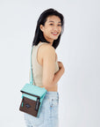 Close up view of a dark haired model facing away from the camera and smiling over her shoulder. She is wearing a tan crop top and jeans. She carries Sherpani crossbody, the Pica in Seagreen, as a crossbody.