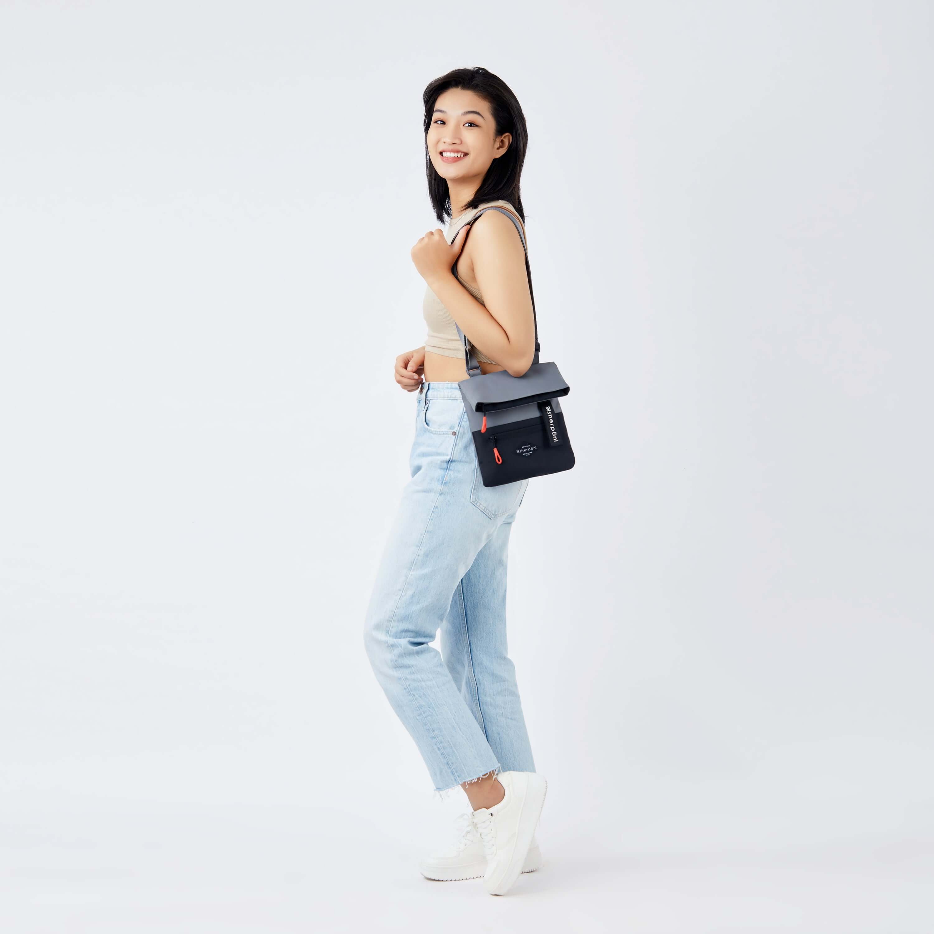 Full body view of a dark hair model facing the side and smiling over her left shoulder. She is wearing a tan crop top, jeans and white sneakers. She carries Sherpani crossbody, the Pica in Stone, over her shoulder. 