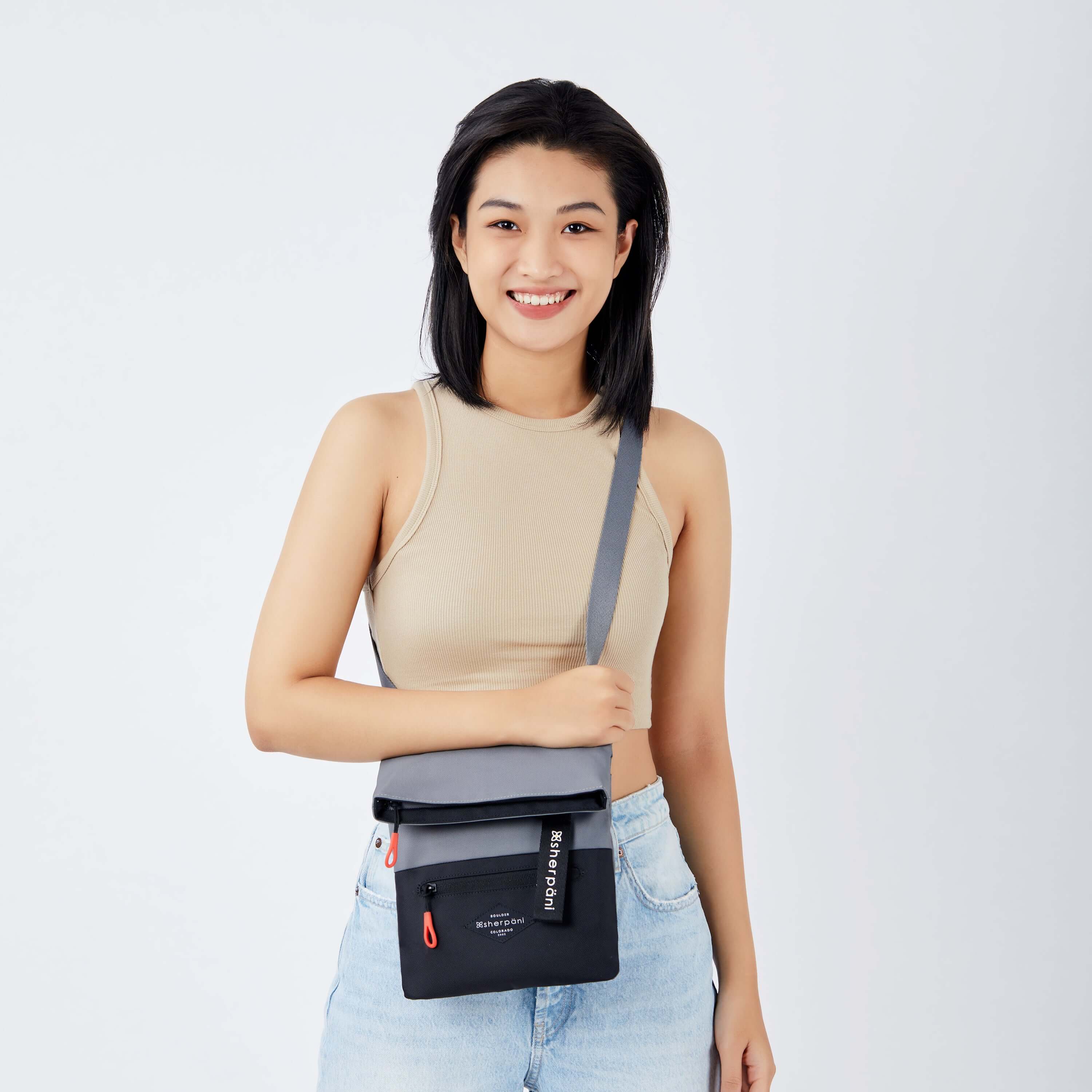 Close up view of a dark haired model facing the camera and smiling. She is wearing a tan crop top and jeans. She carries Sherpani crossbody, the Pica in Stone, as a crossbody. #color_stone