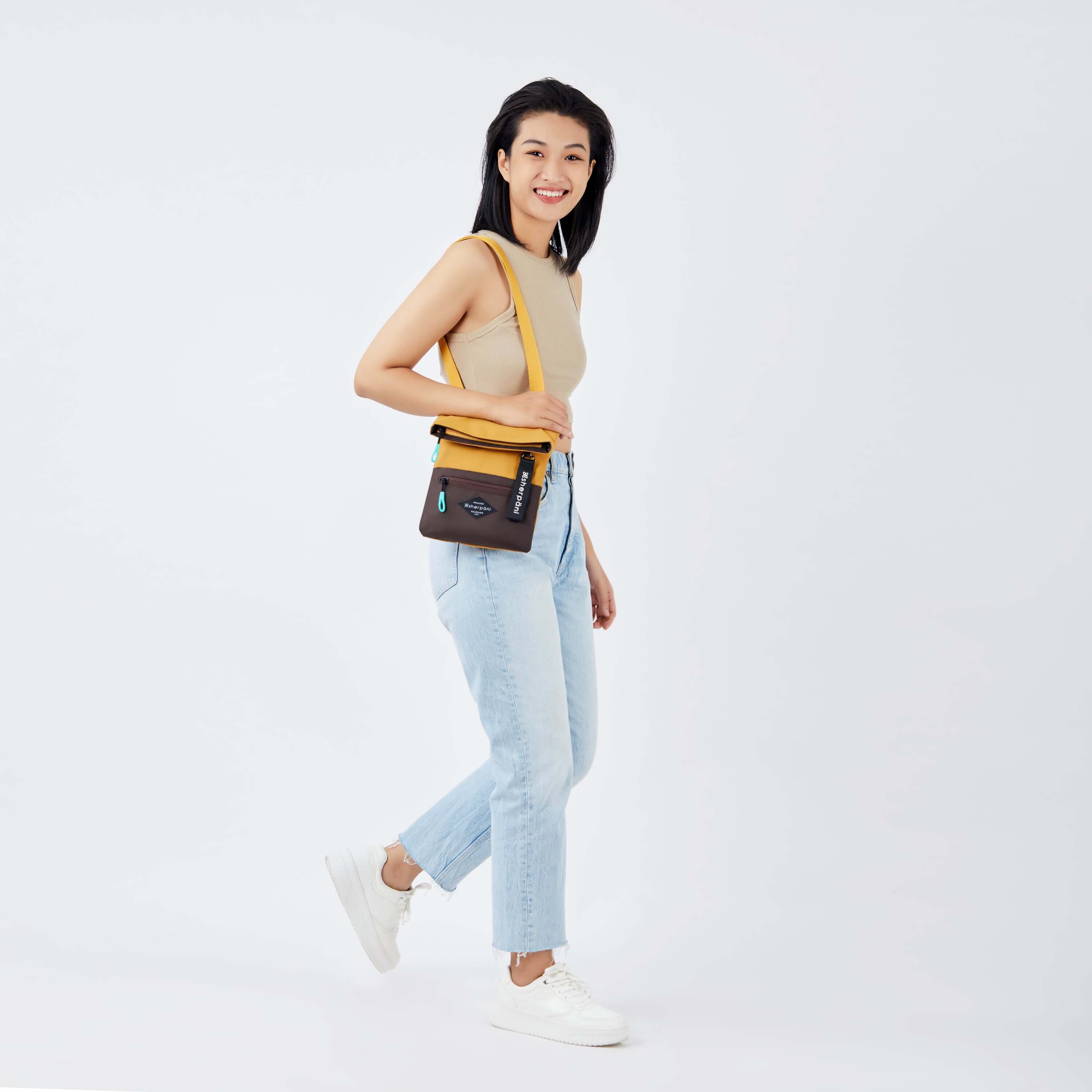 Full body view of a dark haired model facing the side and smiling at the camera. She is wearing a tan crop top, jeans and white sneakers. She carries Sherpani crossbody, the Pica in Sundial, over her shoulder. 