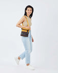 Full body view of a dark haired model facing the side and smiling at the camera. She is wearing a tan crop top, jeans and white sneakers. She carries Sherpani crossbody, the Pica in Sundial, over her shoulder.