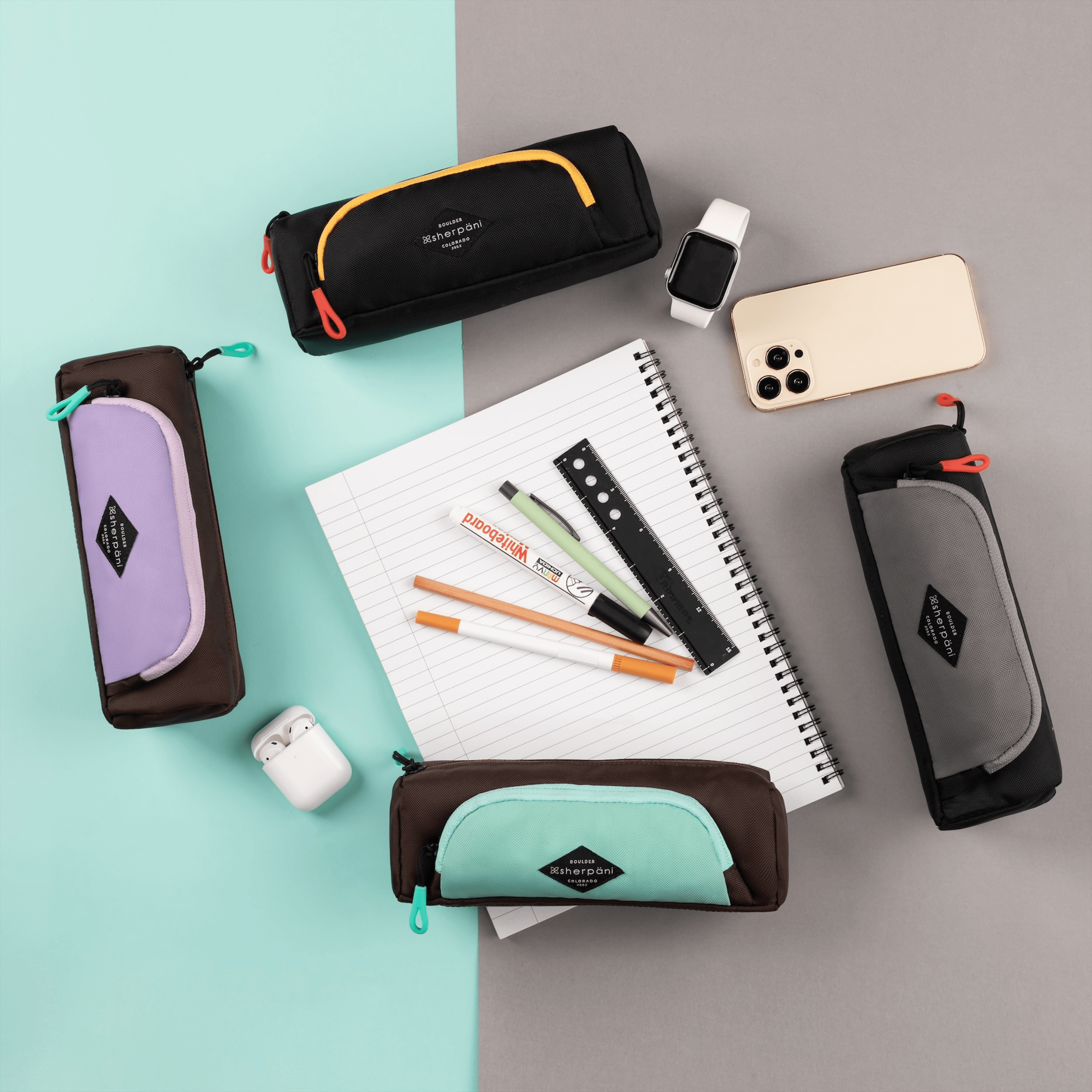 Top view of four Shepani travel accessories arranged in a circle along with example items for their use. Everything lies on a green and gray backdrop. Clockwise from upper left: Poet in Chromatic, smart watch, phone, Jolie in Stone, Jolie in Seagreen, AirPods, Jolie in Lavender. In the center lies a notebook and markers.