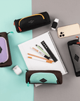 Top view of four Shepani travel accessories arranged in a circle along with example items for their use. Everything lies on a green and gray backdrop. Clockwise from upper left: Poet in Chromatic, smart watch, phone, Jolie in Stone, Jolie in Seagreen, AirPods, Jolie in Lavender. In the center lies a notebook and markers.