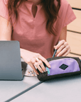 A red haired woman sits in front of a laptop at an outdoor table. She is wearing a pink top. She is pulling a pen out of Sherpani travel accessory, the Poet in Lavender.