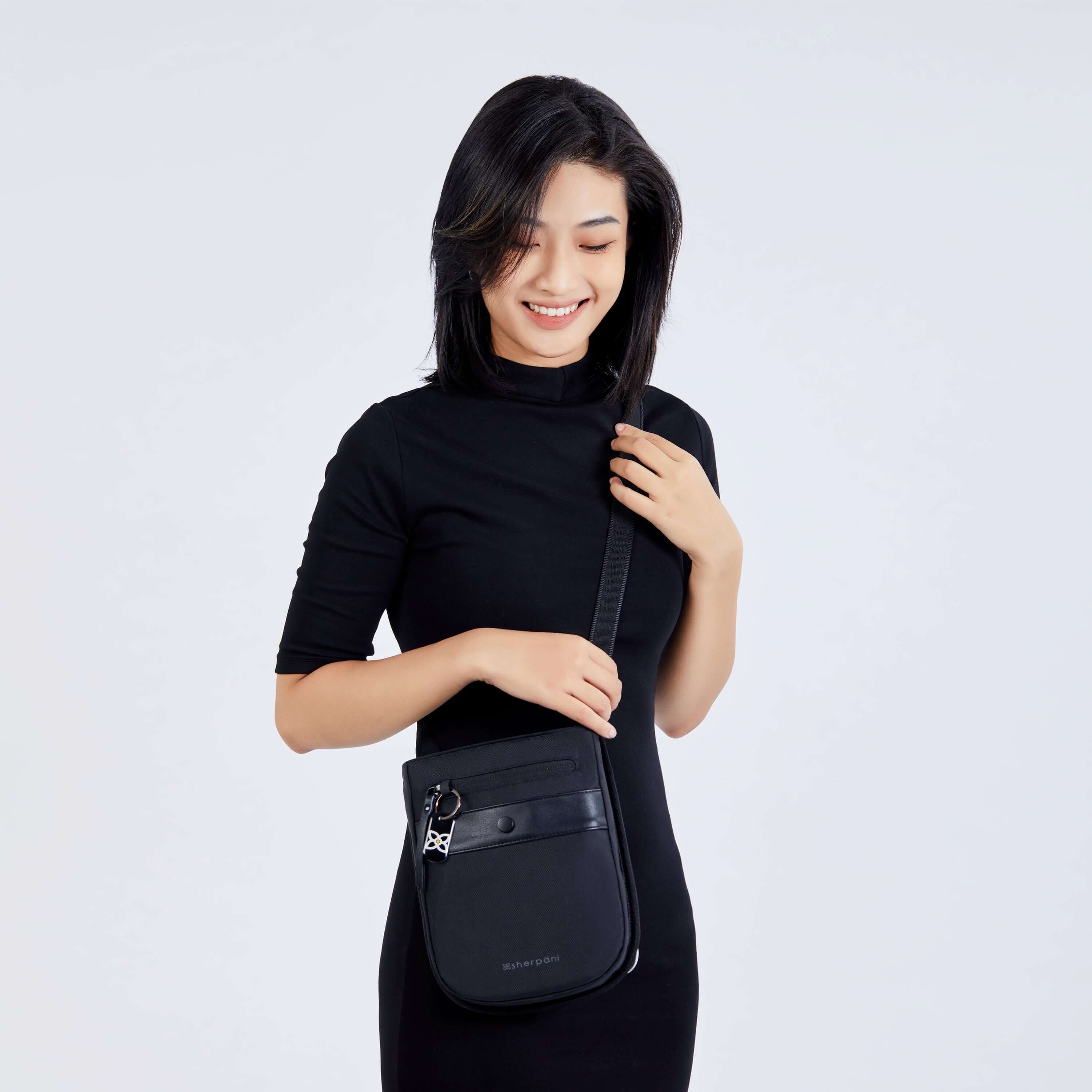 Close up view of a dark haired model smiling down at her bag. She is wearing a black dress and Sherpani's Anti-Theft crossbody the Prima AT in Carbon.